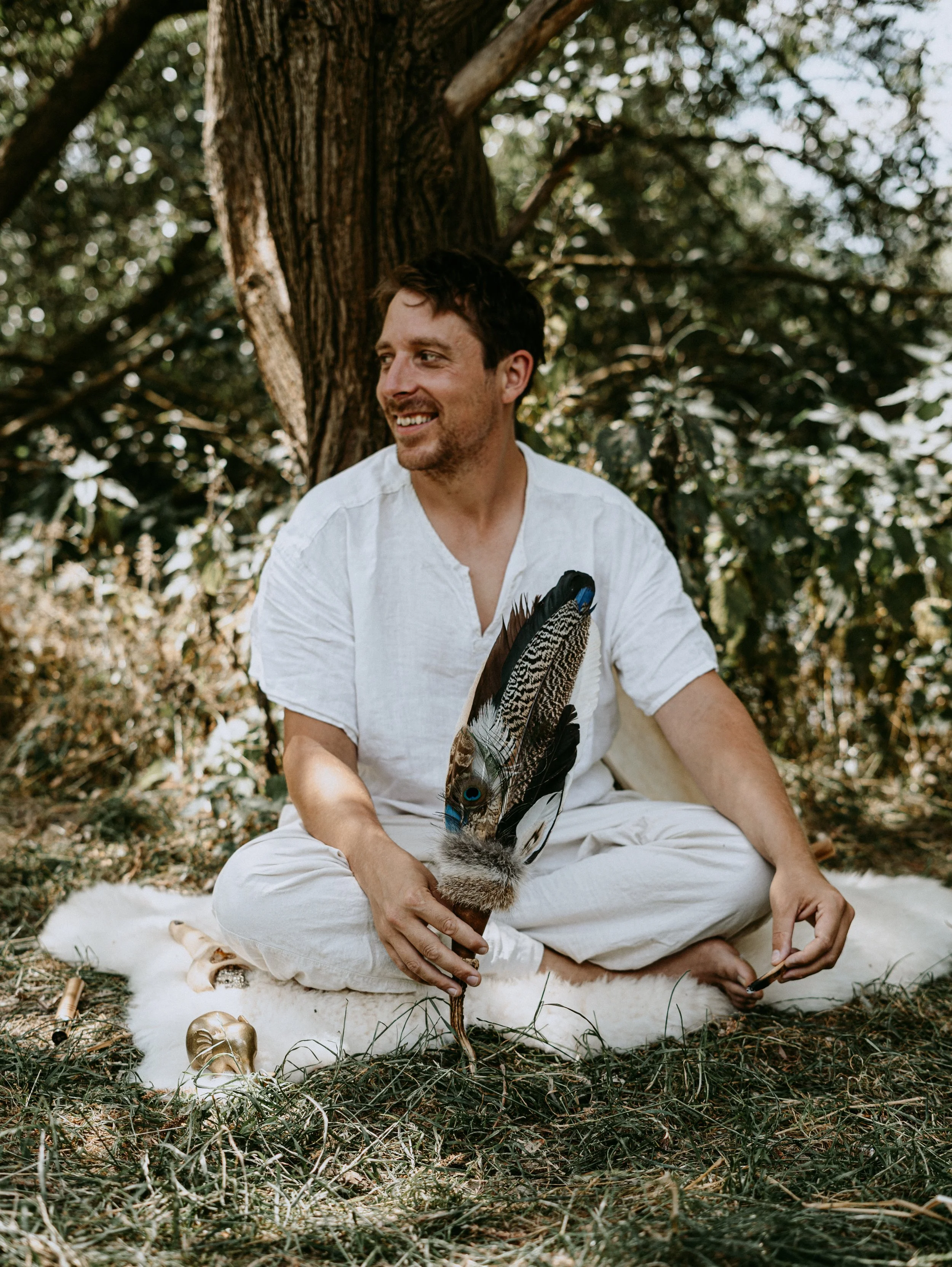 Muž sedící venku se zkříženýma nohama na bílém kožešinovém koberci pod stromem, držící velký opeřený vějíř s pavími pery, usmívající se a odvracející pohled, s tmavě zbarveným ptákem na zemi poblíž.