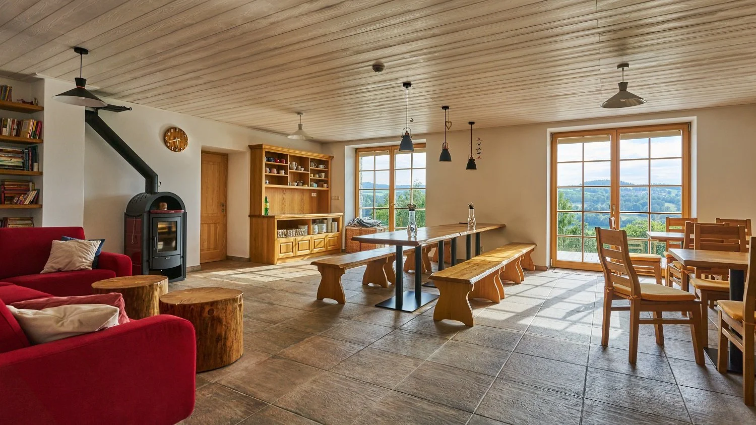 Living room with wooden ceiling, large windows showing outdoor landscape, red couch, wooden coffee tables, wood-paneled door, bookshelf, wood stove, dining table with benches, and additional chairs.