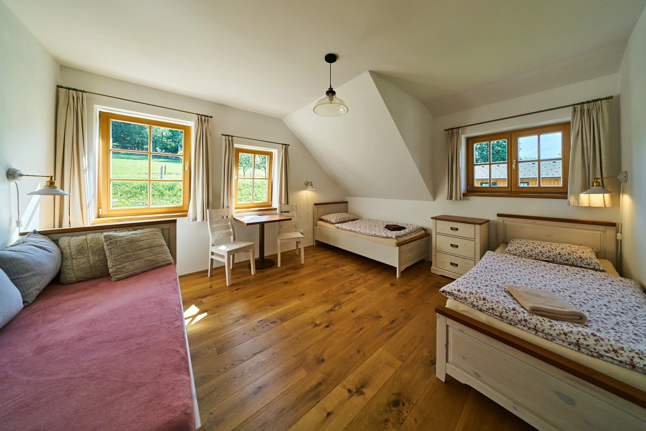 A cozy bedroom with three windows showing a green outdoor landscape, wooden floors, two beds with floral bedspreads, and a sitting area with a table and chairs.