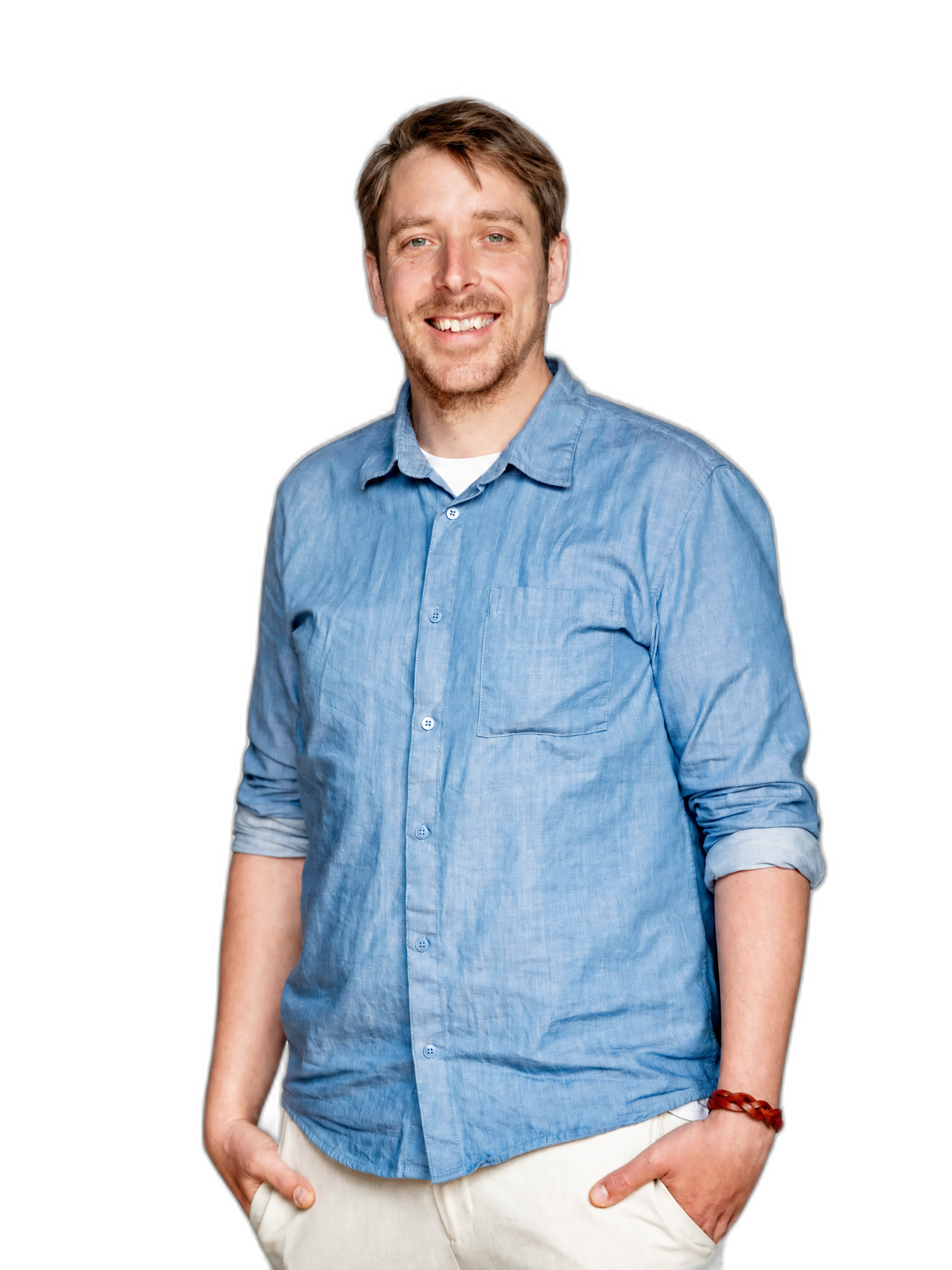 A smiling young man with light brown hair wearing a light blue button-up shirt with sleeves rolled up, beige pants, and a red bracelet on his left wrist, standing against a black background.