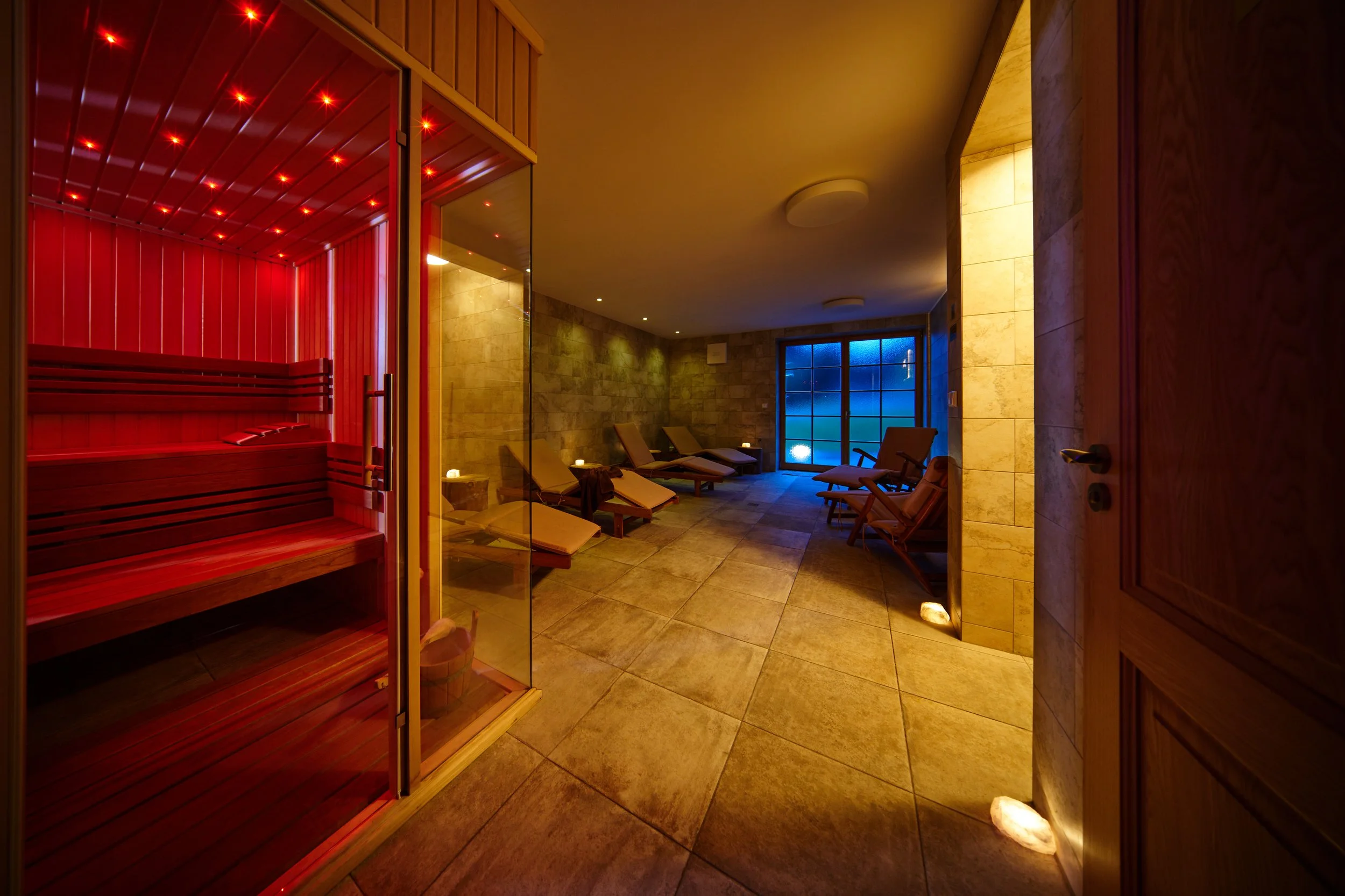 A relaxation room featuring a wooden sauna with red lighting on the left, lounge chairs on the right, and a window with blue light in the background.