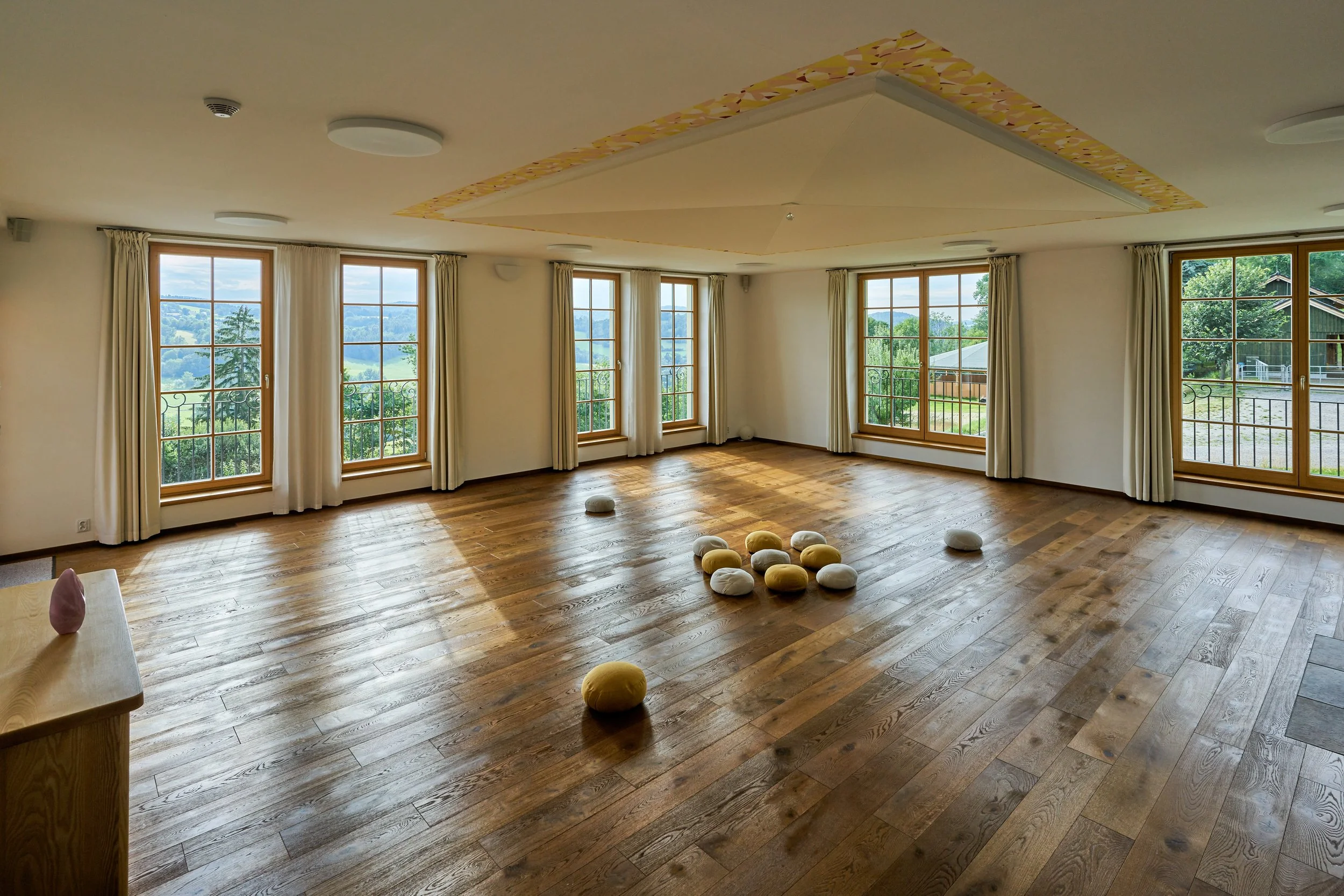 A spacious room with large windows showing a scenic landscape, hardwood floors, cream-colored curtains, and a ceiling feature with decorative trim. Several cushions are scattered on the floor.