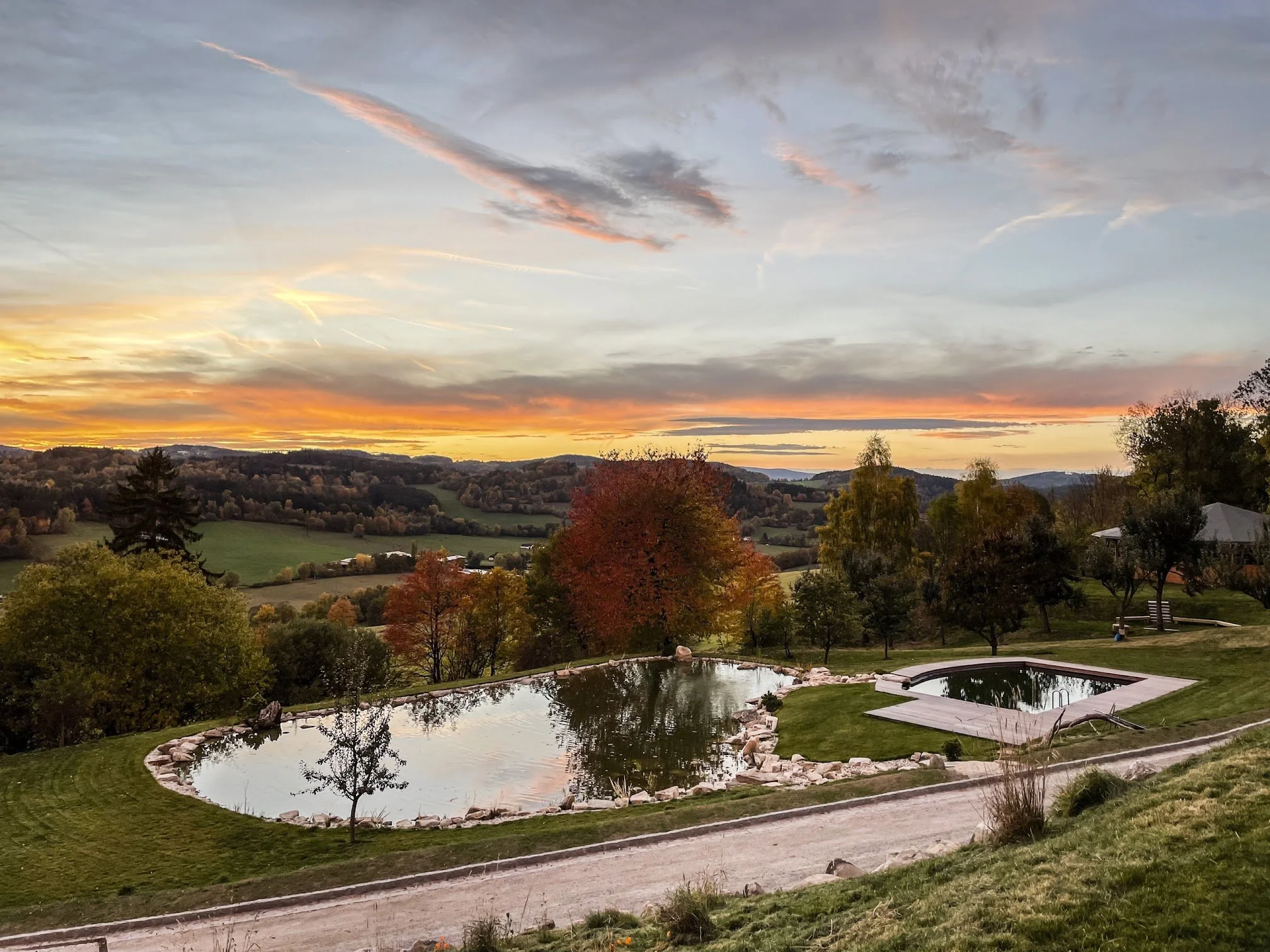 Scenic view of a sunset over rolling hills with trees in autumn colors, a small pond with a stone border, and a building with a curved roof in the foreground.