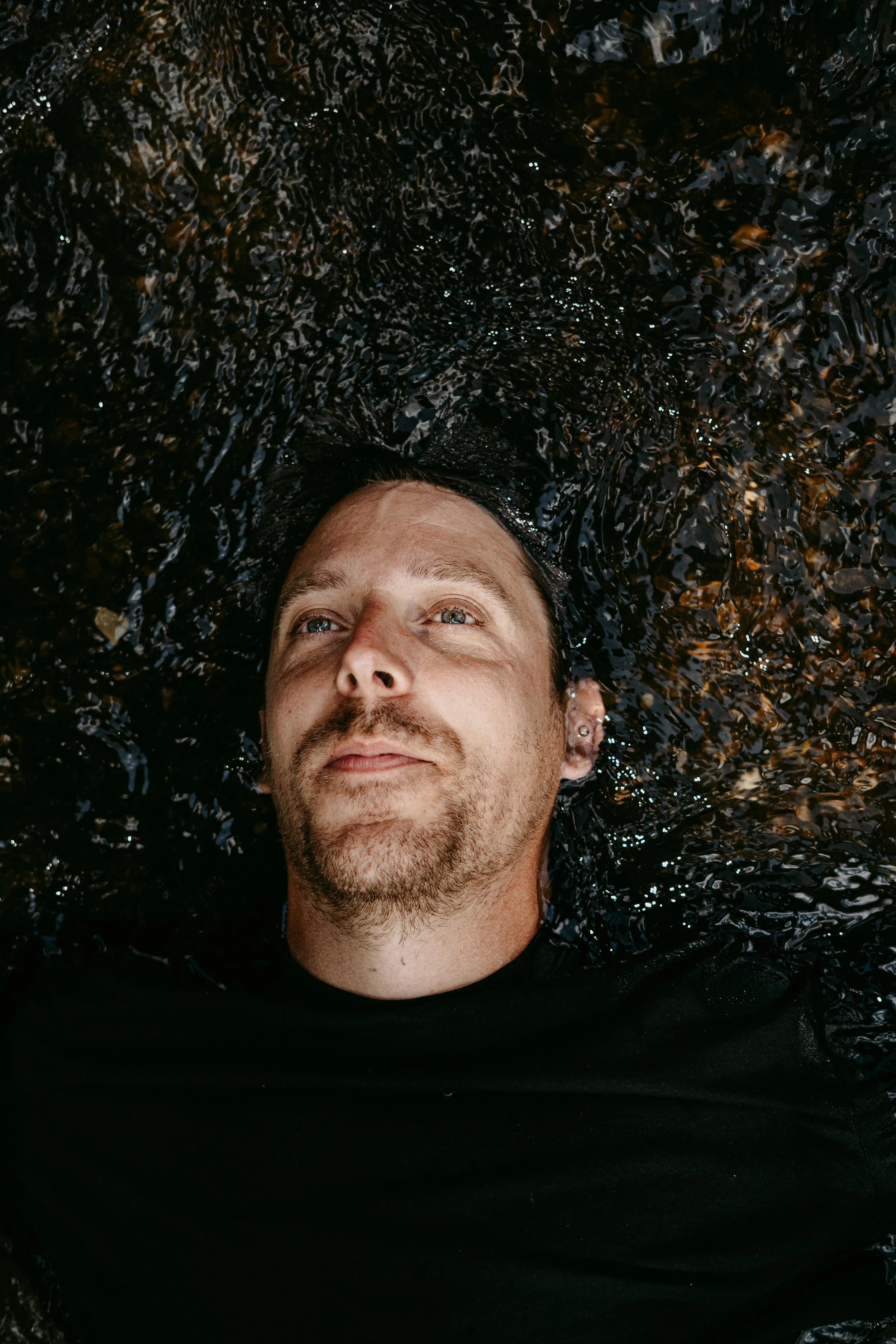 A man lying in dark water, facing upwards with his eyes open.