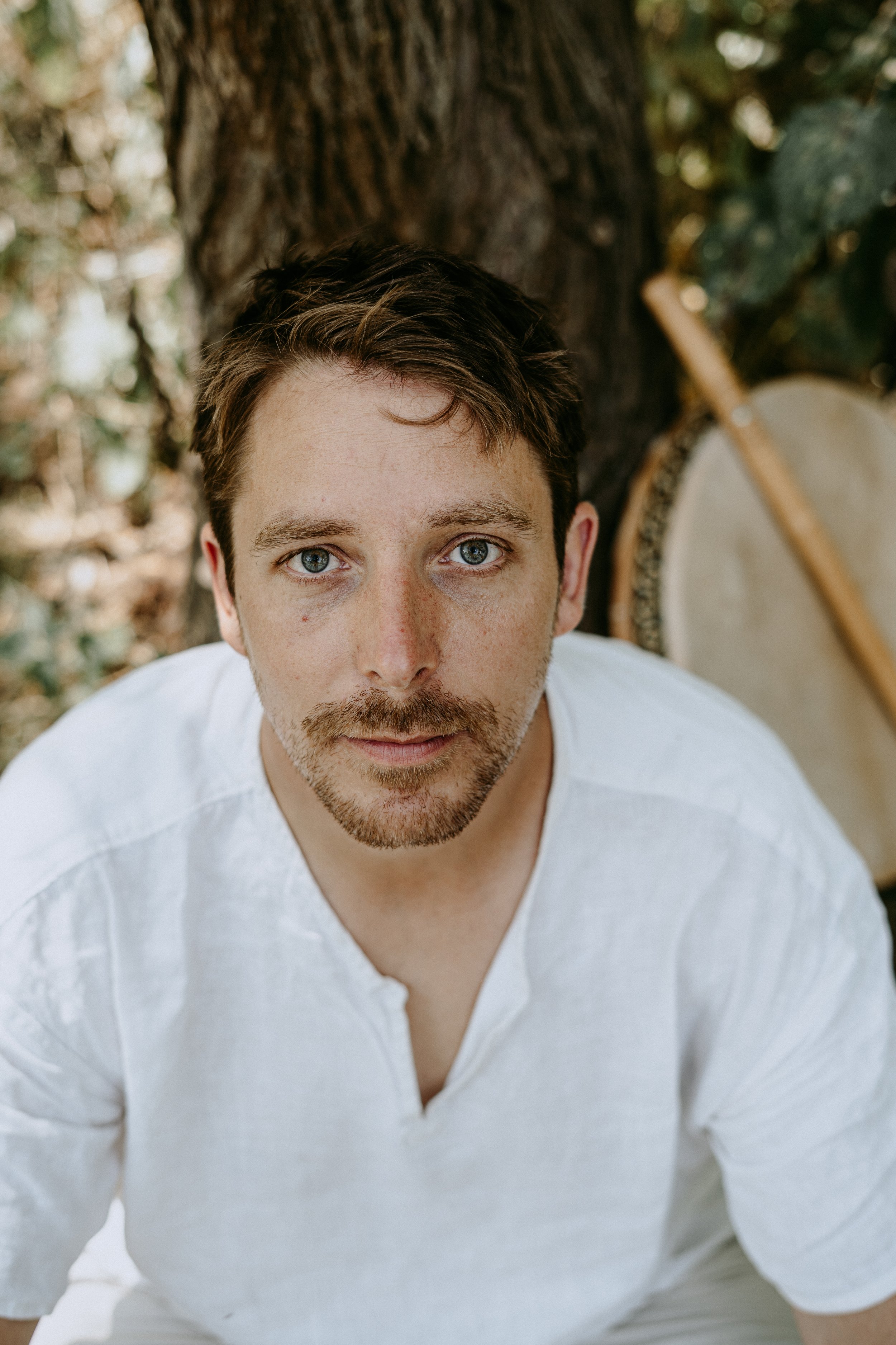 A man with short brown hair, blue eyes, and a beard, is sitting outdoors against a tree, wearing a white shirt. There is a drum and a drumstick in the background.