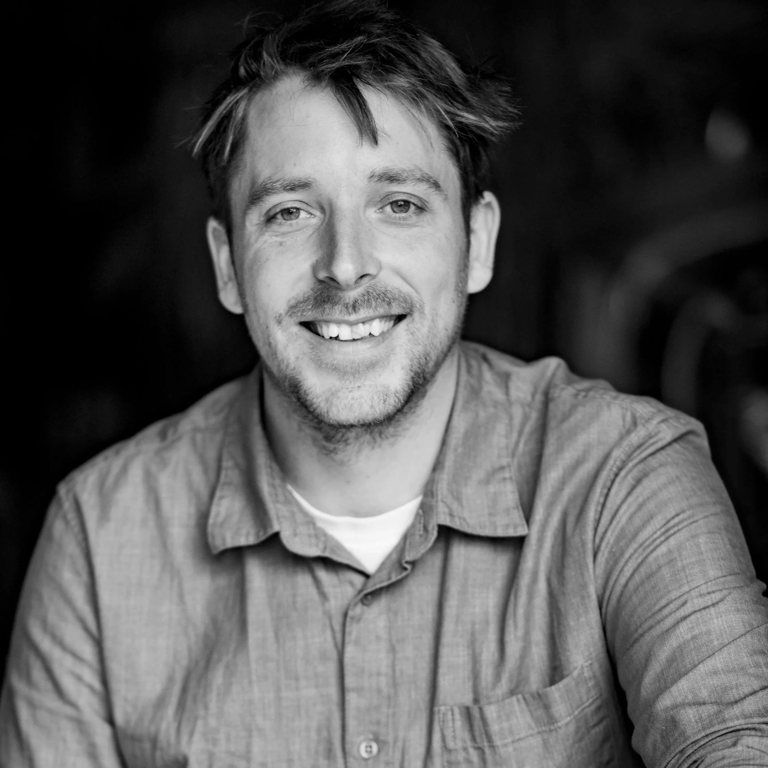 Black and white close-up photograph of a man with short, tousled hair, smiling and wearing a light-colored, collared shirt with vertical stripes.