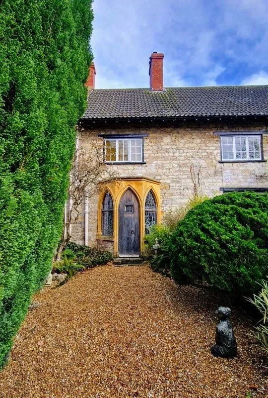 The front entrance of a stone house with a black door, surrounded by a gravel pathway, greenery, and a small statue.
