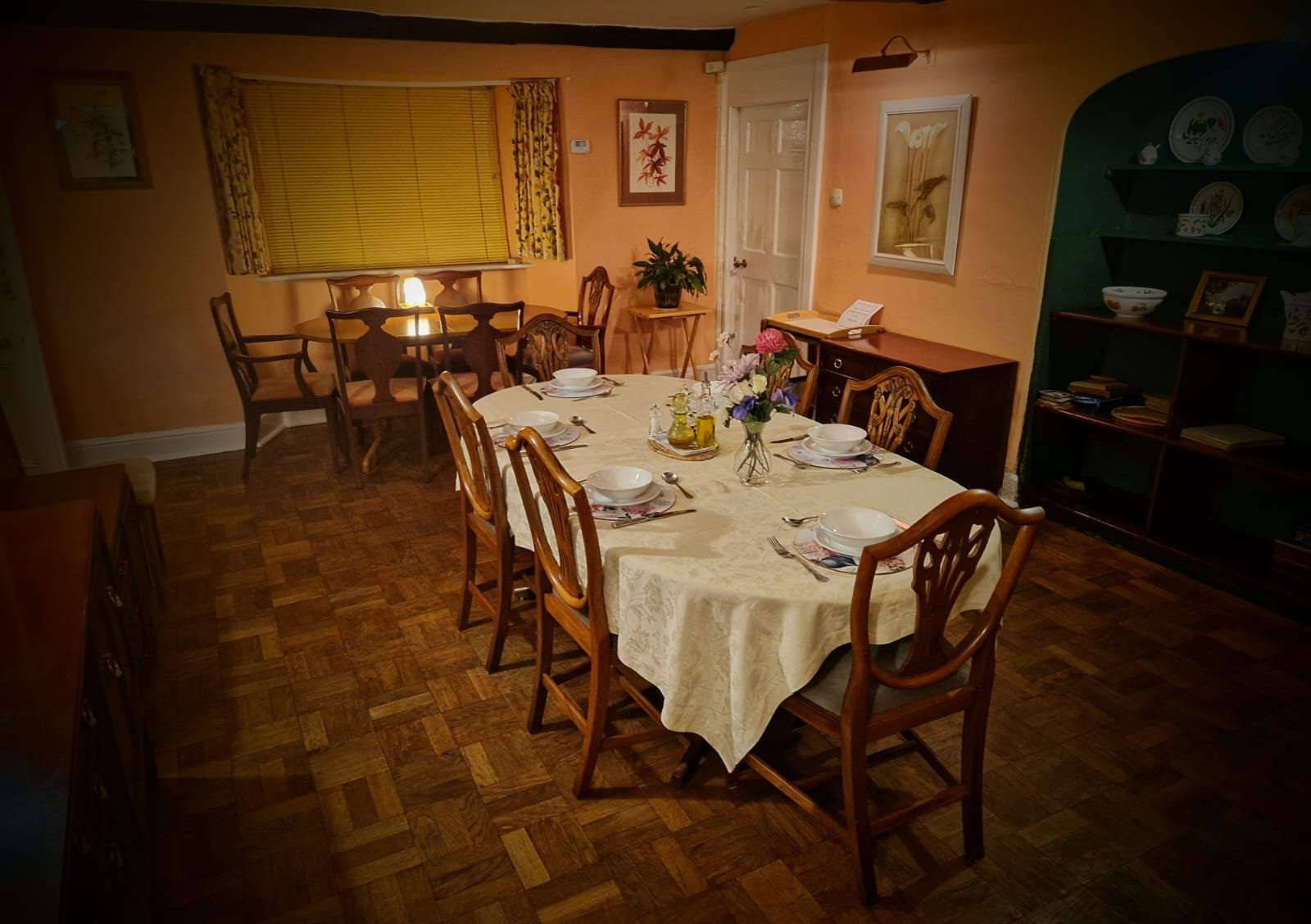 Dining room with a large oval table set with bowls, plates, utensils, a flower centerpiece, and a tablecloth. There are wooden chairs around the table, a window with yellow blinds, artwork on the walls, a sideboard, a bookshelf, and a small lamp prov