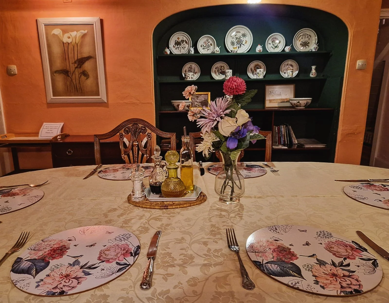 A dining table set for six, with floral placemats, silverware, and a centerpiece of fresh flowers in a glass vase. Behind the table, a black shelf with decorative plates, bowls, and framed artwork is visible, along with a framed painting of flowers o