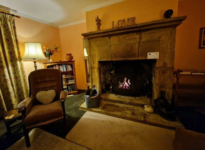 Cozy living room with a lit fireplace, a brown armchair with a heart-shaped cushion, a side table, a bookshelf, a table lamp, and decorative items on the mantle including a sculpture spelling 'LOVE' and a small vase. A basket and some books are on th