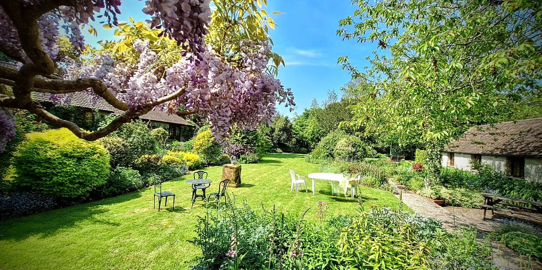 A lush, vibrant garden with blooming trees including a large tree with purple flowers, surrounded by green shrubs and flowering plants. There are outdoor chairs and a table, a stone pathway, and a building with a tiled roof in the background under a 