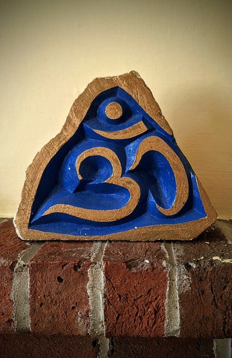 A decorative wooden sculpture in the shape of a triangle with carved blue and gold spiritual symbols, placed on a brick surface against a plain wall background.