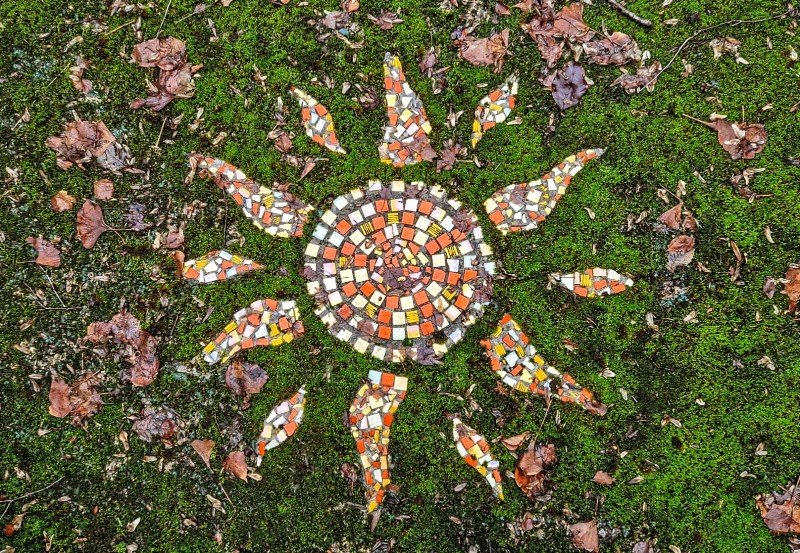 Mosaic sun design on mossy ground surrounded by fallen leaves.