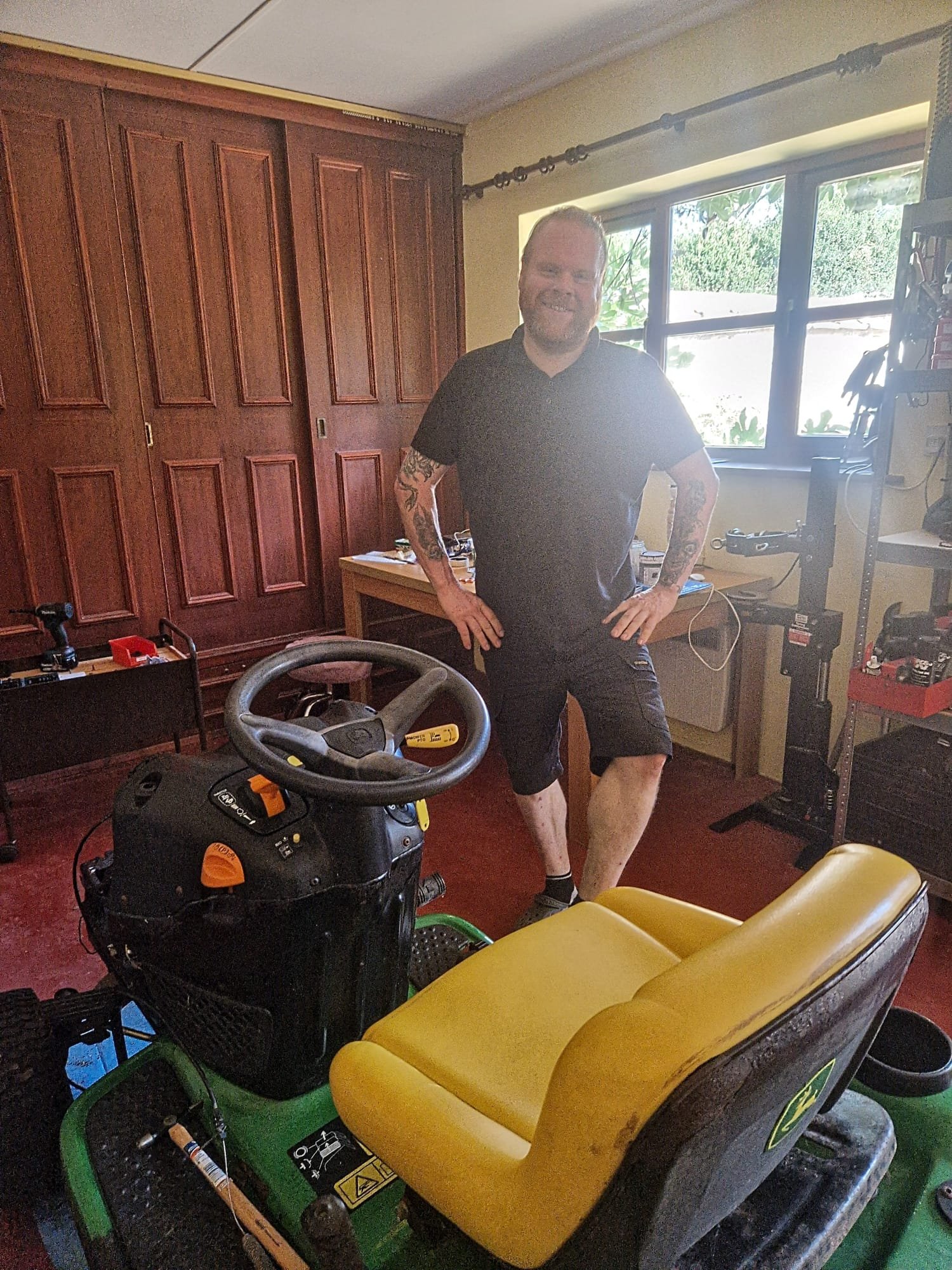 Man standing indoors near large window, smiling, wearing black t-shirt and shorts, tattoos on arms, surrounded by various tools and equipment including a yellow wheelbarrow and a shop vacuum.