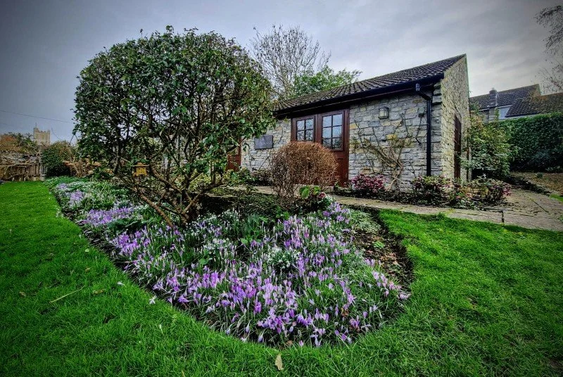 A small stone house surrounded by a lush garden with colorful flowers, a single bush, and a well-maintained lawn.
