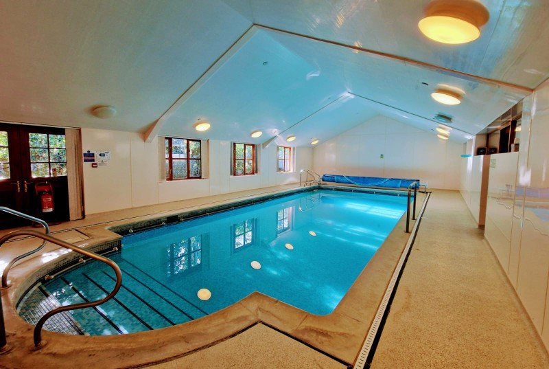 Indoor swimming pool with a blue cover rolled up at the far end, large windows letting in natural light, and ceiling lights reflecting on the water, with a handrail and steps leading into the pool.