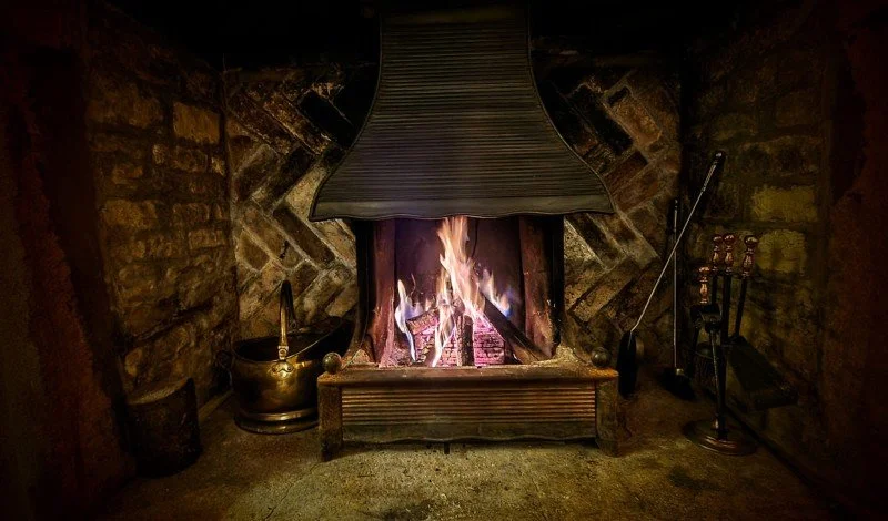 Indoor rustic fireplace with a fire burning, surrounded by brick walls and stone accents, with fireplace tools and a brass kettle nearby.