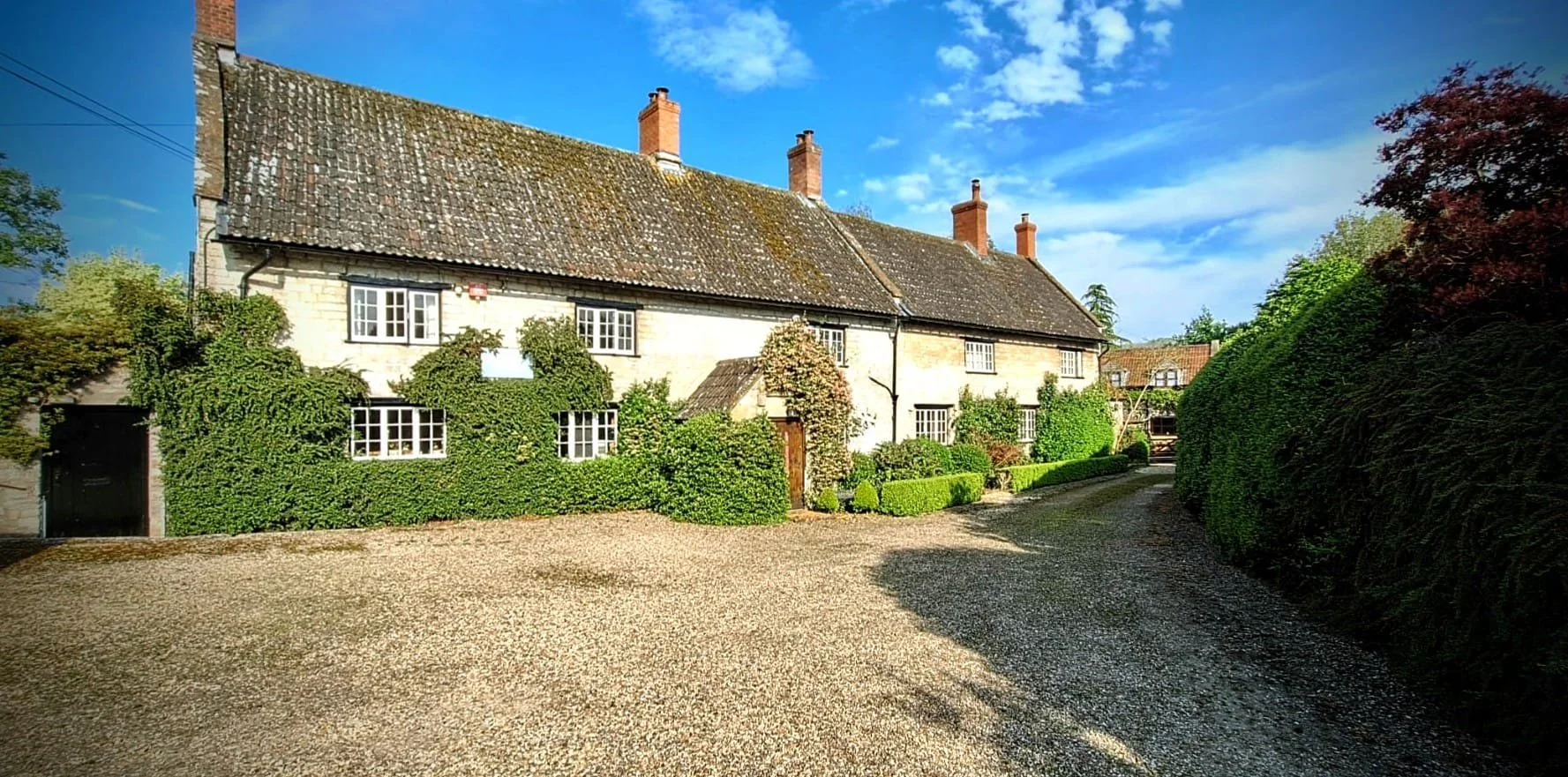 A charming country house with a weathered tiled roof, white walls, and multiple chimneys. The house is surrounded by lush green bushes and trees, with a gravel driveway winding in front of it. Bright blue sky with some clouds overhead.