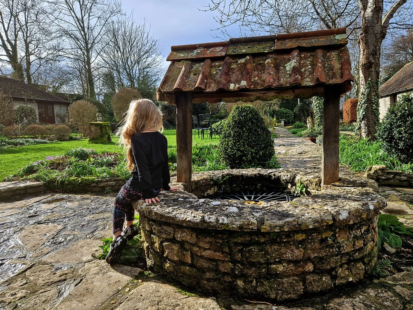 Child with long blonde hair leaning on a stone well in a garden, surrounded by trees and shrubs, with a path and outdoor furniture in the background.