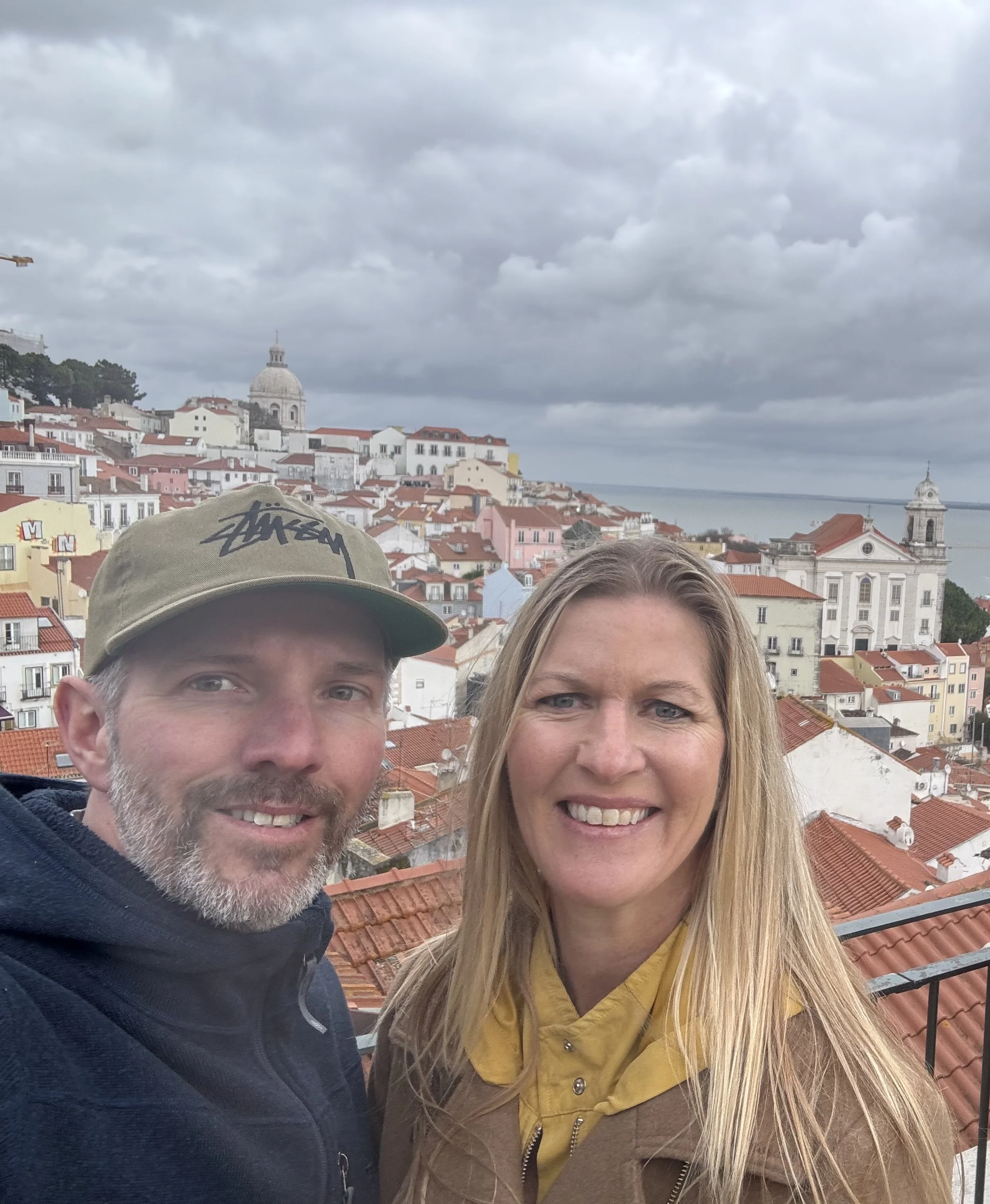 A smiling couple takes a selfie with a colorful cityscape and cloudy sky in the background.