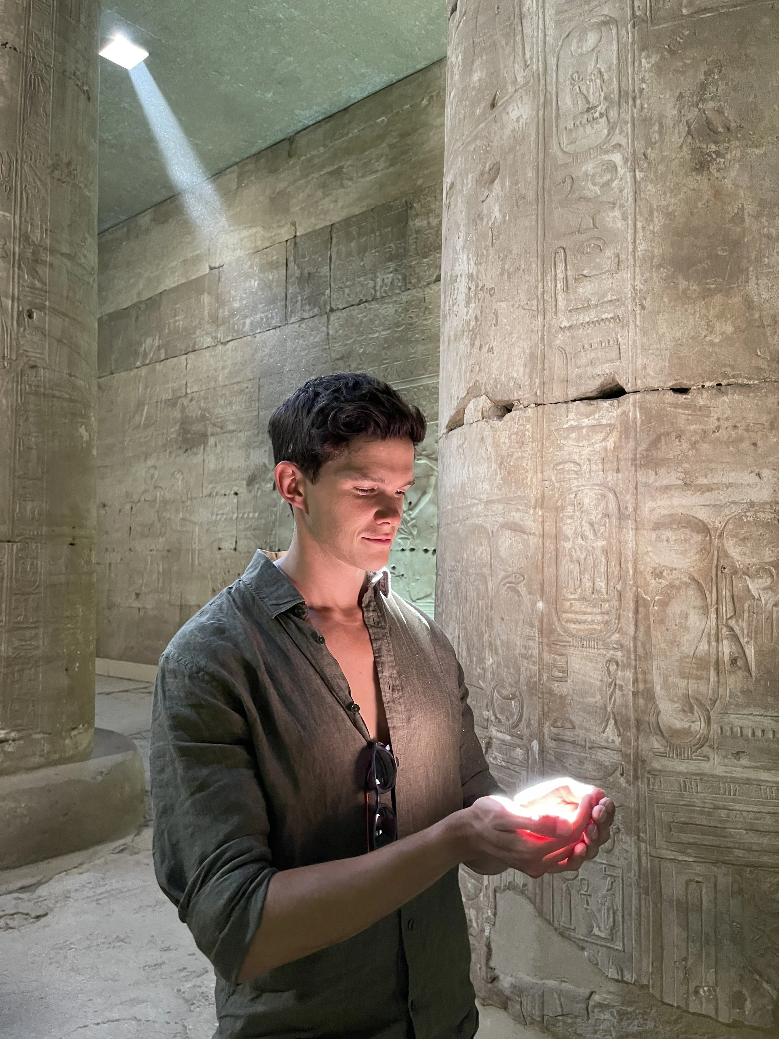 A young man with dark hair, wearing a dark button-up shirt, holds a glowing object in his hands while standing in front of ancient Egyptian hieroglyphic carvings on stone walls inside a historical site.
