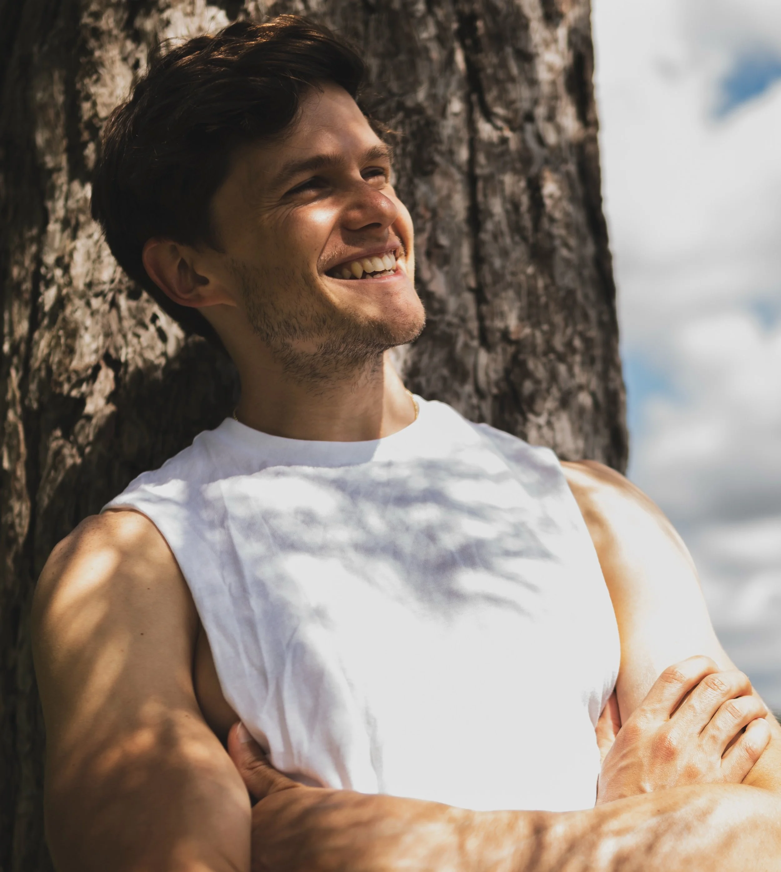 Tom Prior with a big smile, leaning against a tree outdoors on a sunny day.