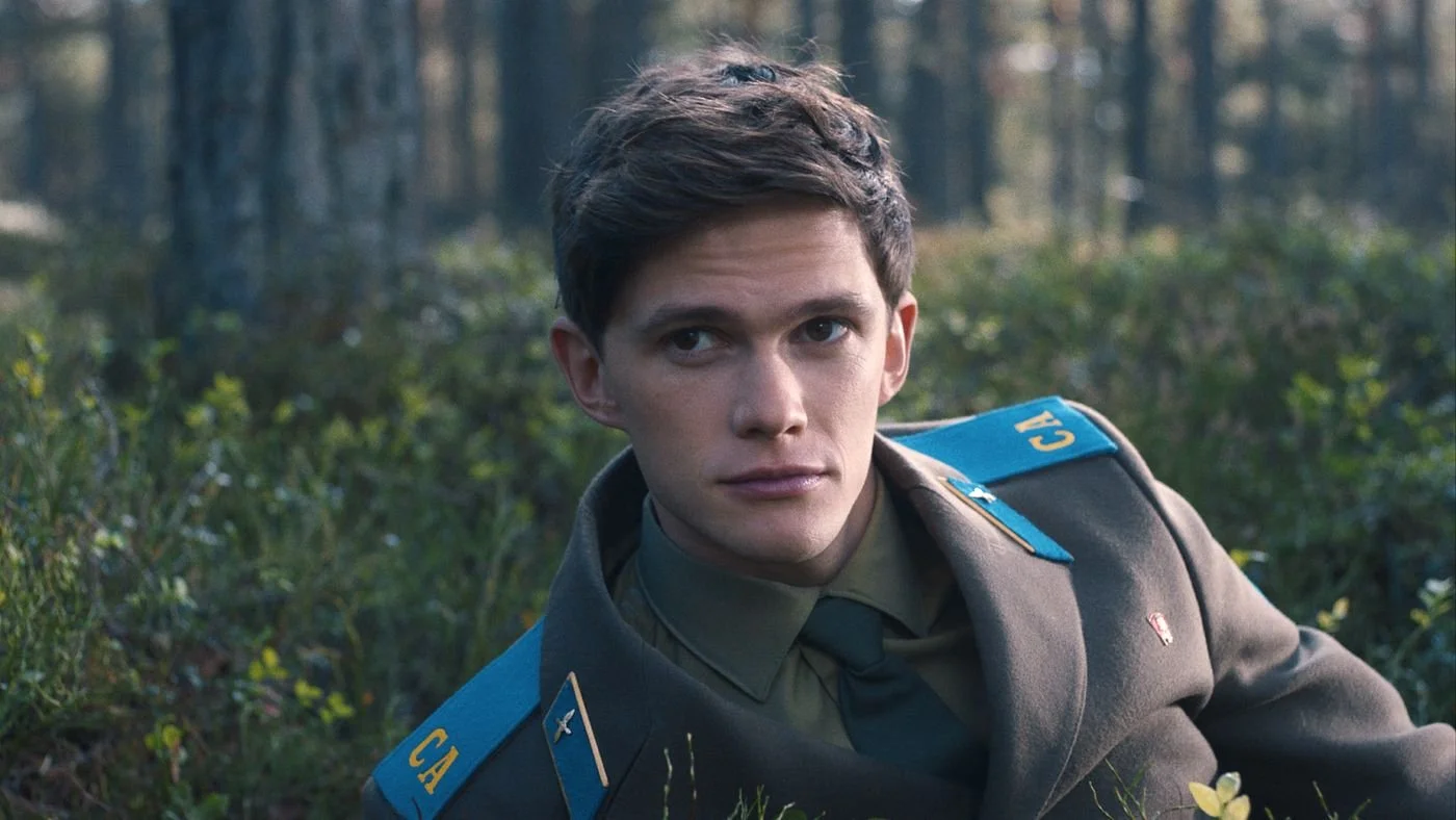 A young man in a military uniform lying on the ground in a forest, looking to the side with a serious expression.
