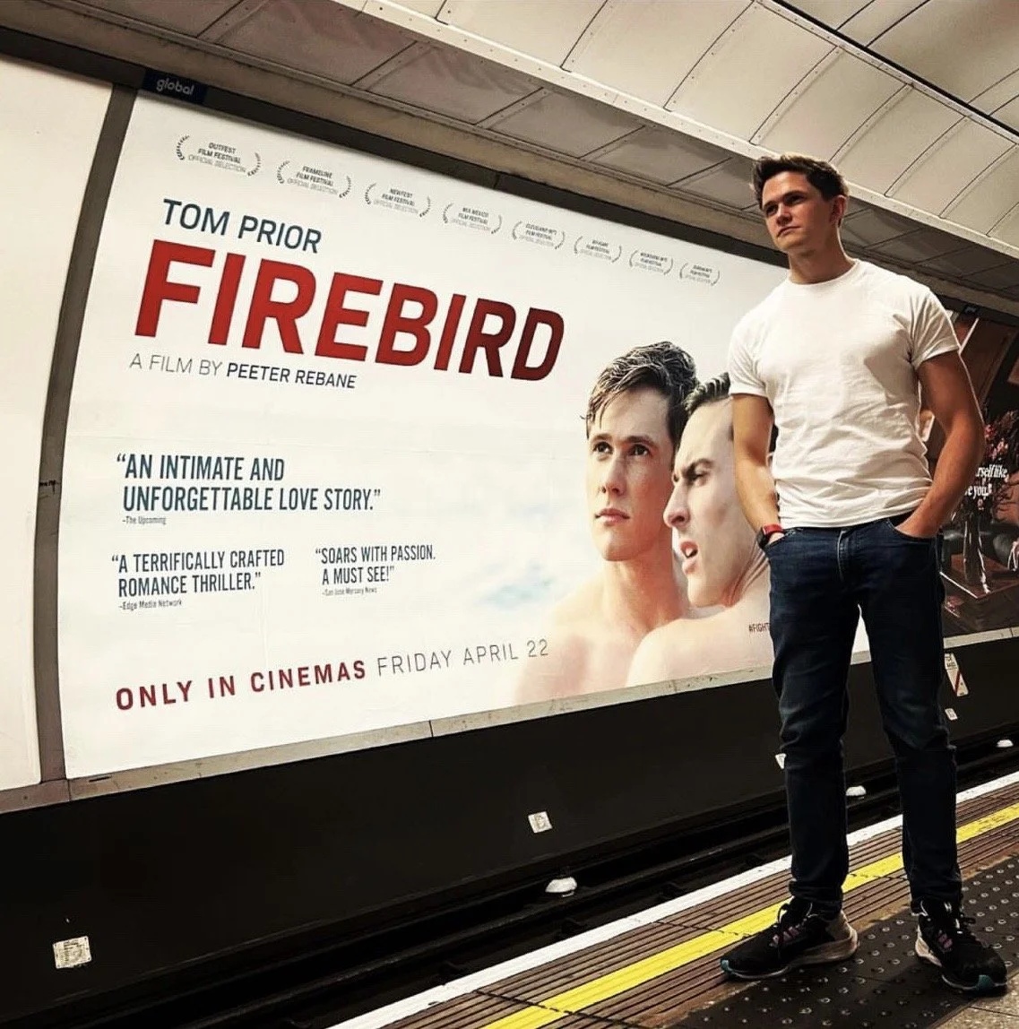 A young man in a white t-shirt and dark jeans standing on a subway platform next to a large movie poster for 'Firebird' featuring two women with serious expressions and film quotes, promoting the film's release on April 22.