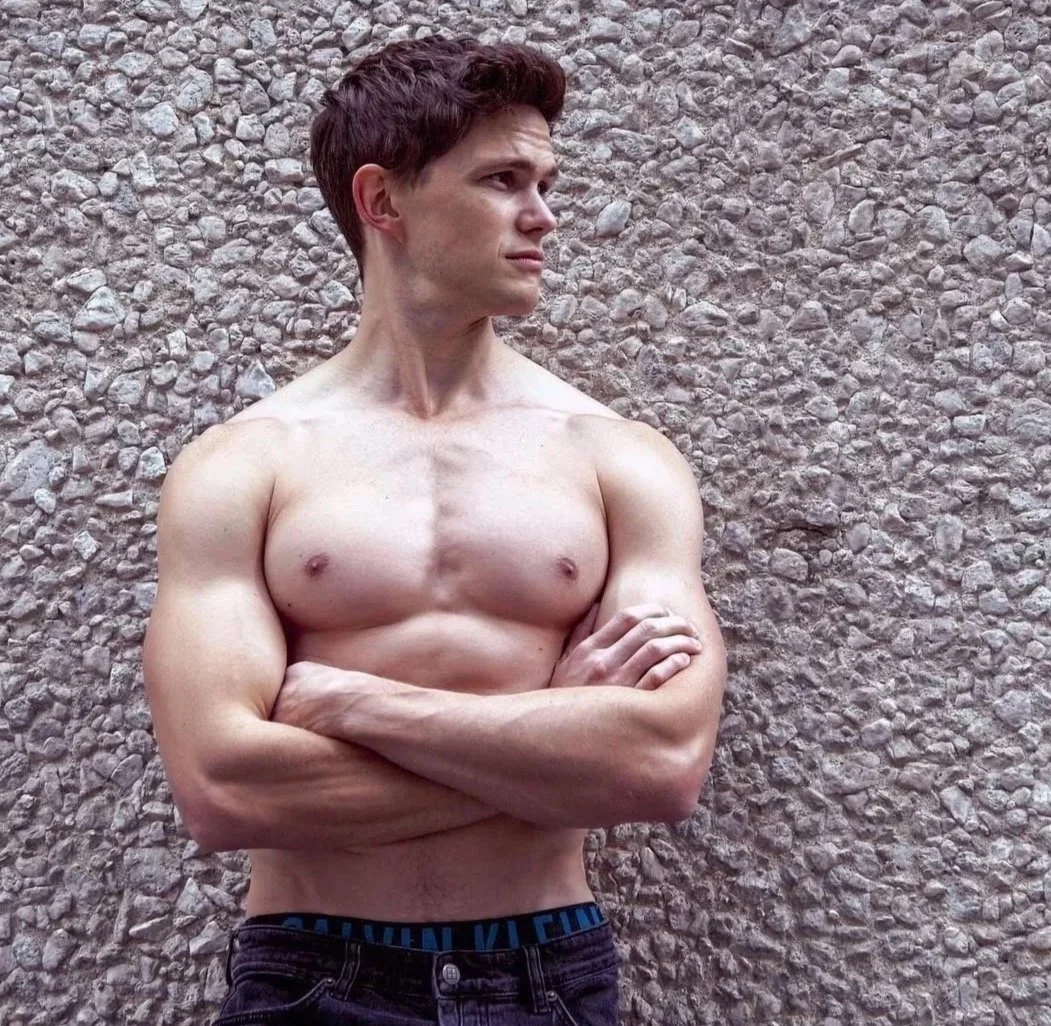 A shirtless young man with crossed arms standing against a textured stone wall, looking to his left.