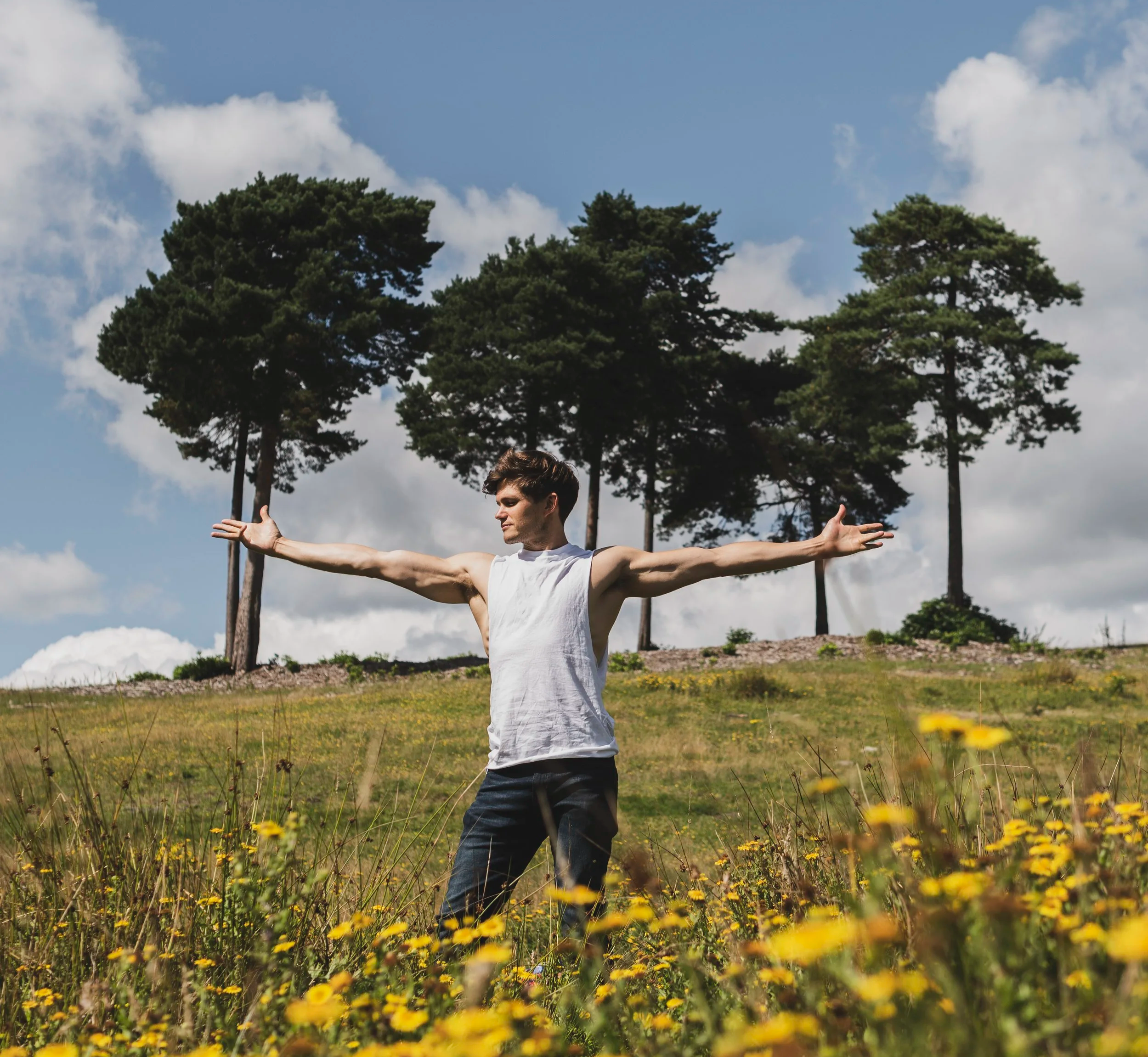 Young man standing in a field of yellow flowers with arms outstretched, trees and cloudy sky in the background.
