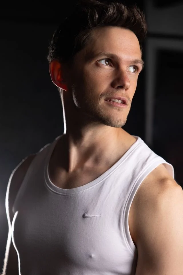 A young man in a white sleeveless athletic shirt looking to the side with earphones in his ears in a gym setting.