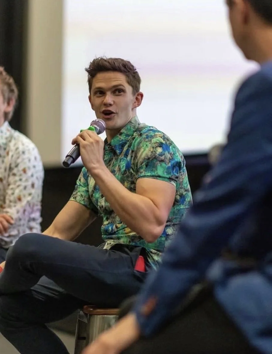 A young man with brown hair wearing a colorful Hawaiian shirt speaking into a microphone during a panel discussion or event, with other people seated around him.