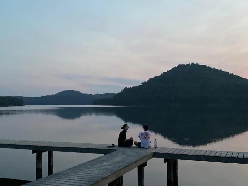Two people sitting on a dock by a calm lake, with hills in the background, during sunset or dusk.
