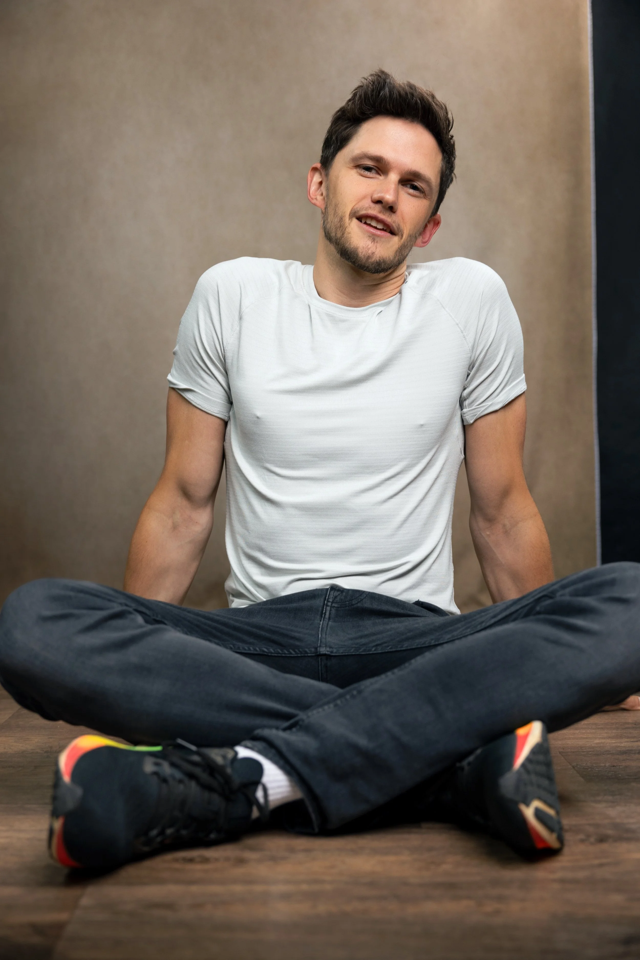 Tom Prior sitting on the floor with his legs crossed, wearing a white t-shirt and black jeans, smiling at the camera.