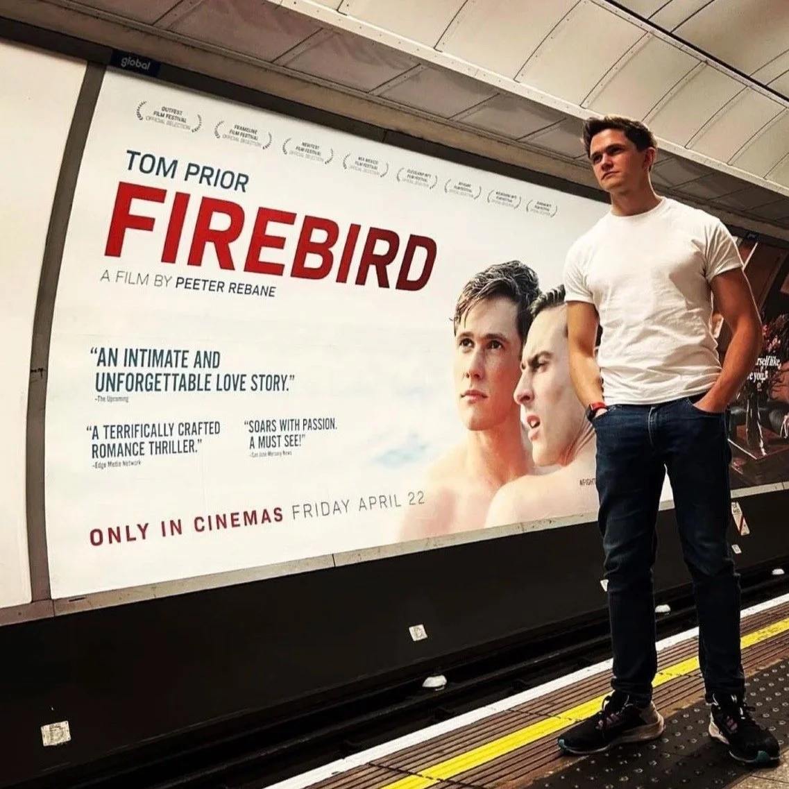 Tom Prior in a white t-shirt and jeans standing on a subway platform next to a large movie poster for 'Firebird' featuring two young men's faces.