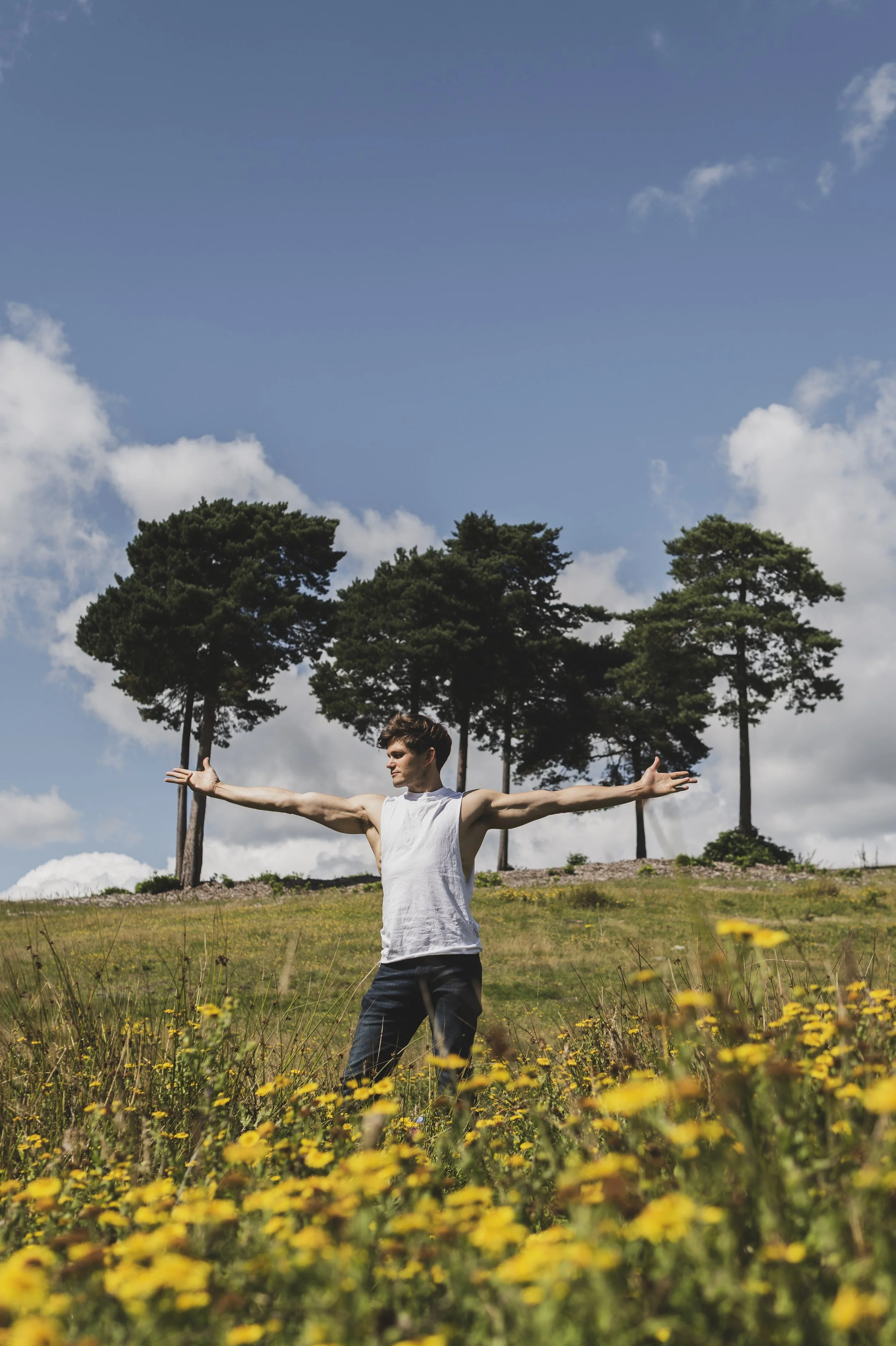 Tom Prior standing in a field of yellow flowers with his arms outstretched, surrounded by trees and under a partly cloudy sky.