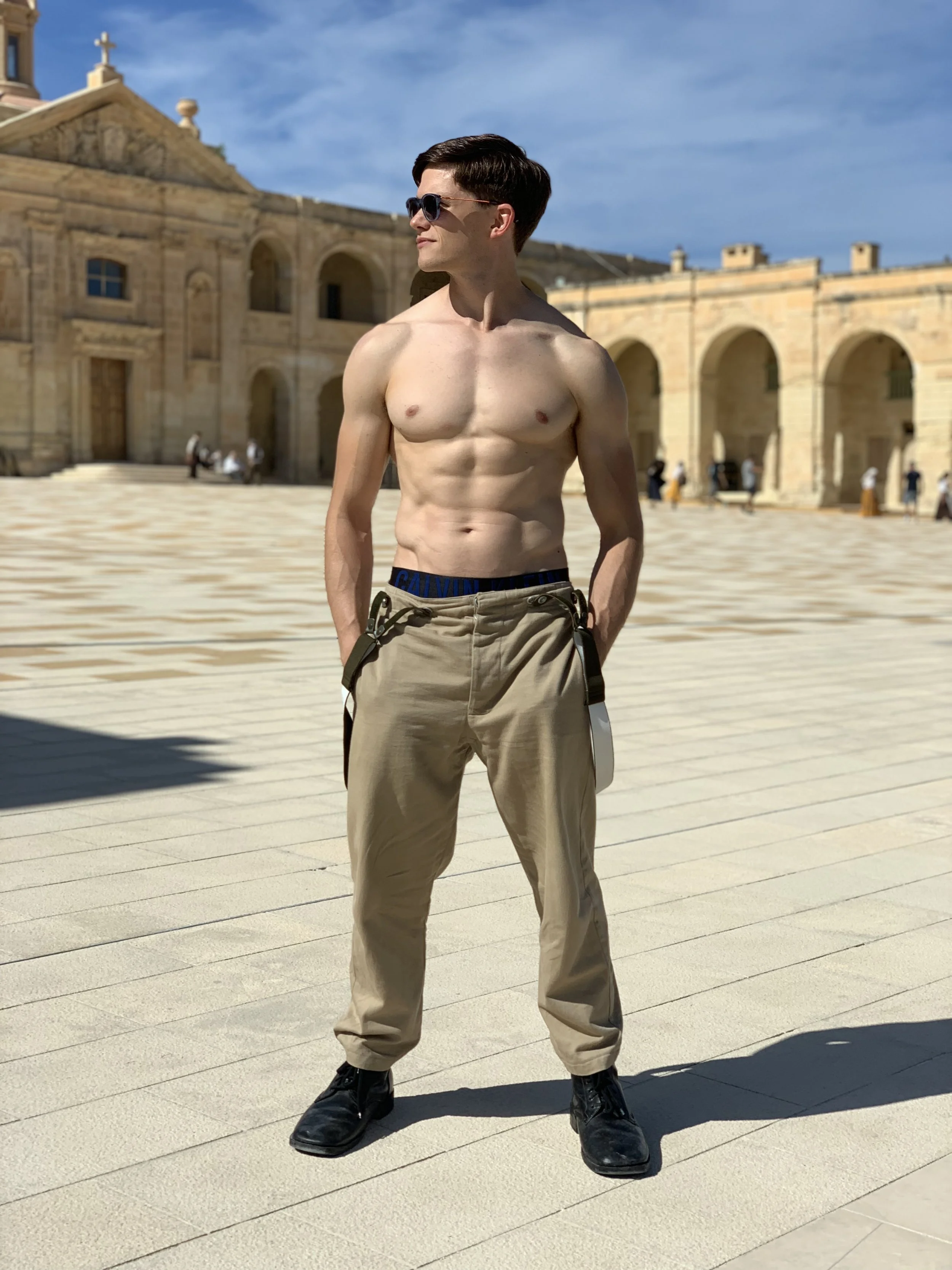 Young man with short dark hair, wearing sunglasses, beige cargo pants, black boots, and no shirt, standing outdoors on a sunny day.
