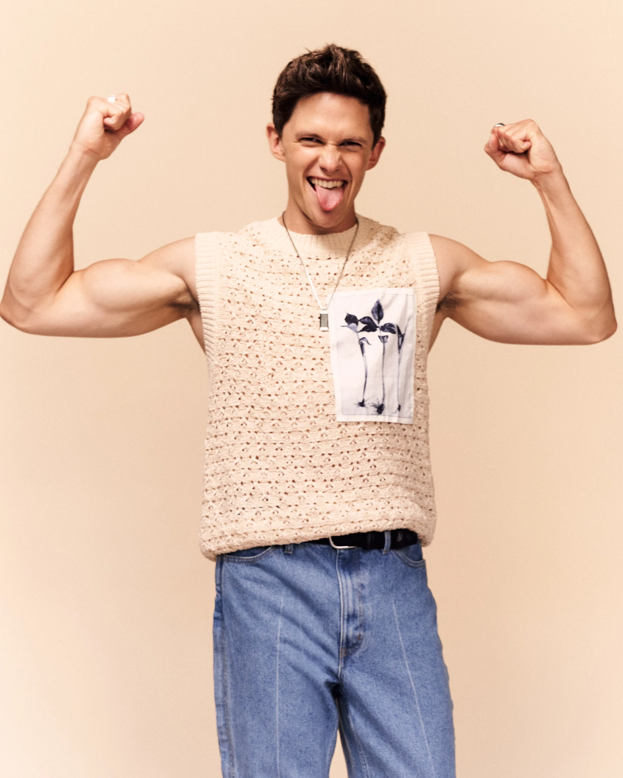 Young man flexing biceps with tongue out wearing sleeveless knitted sweater and jeans against plain beige background.