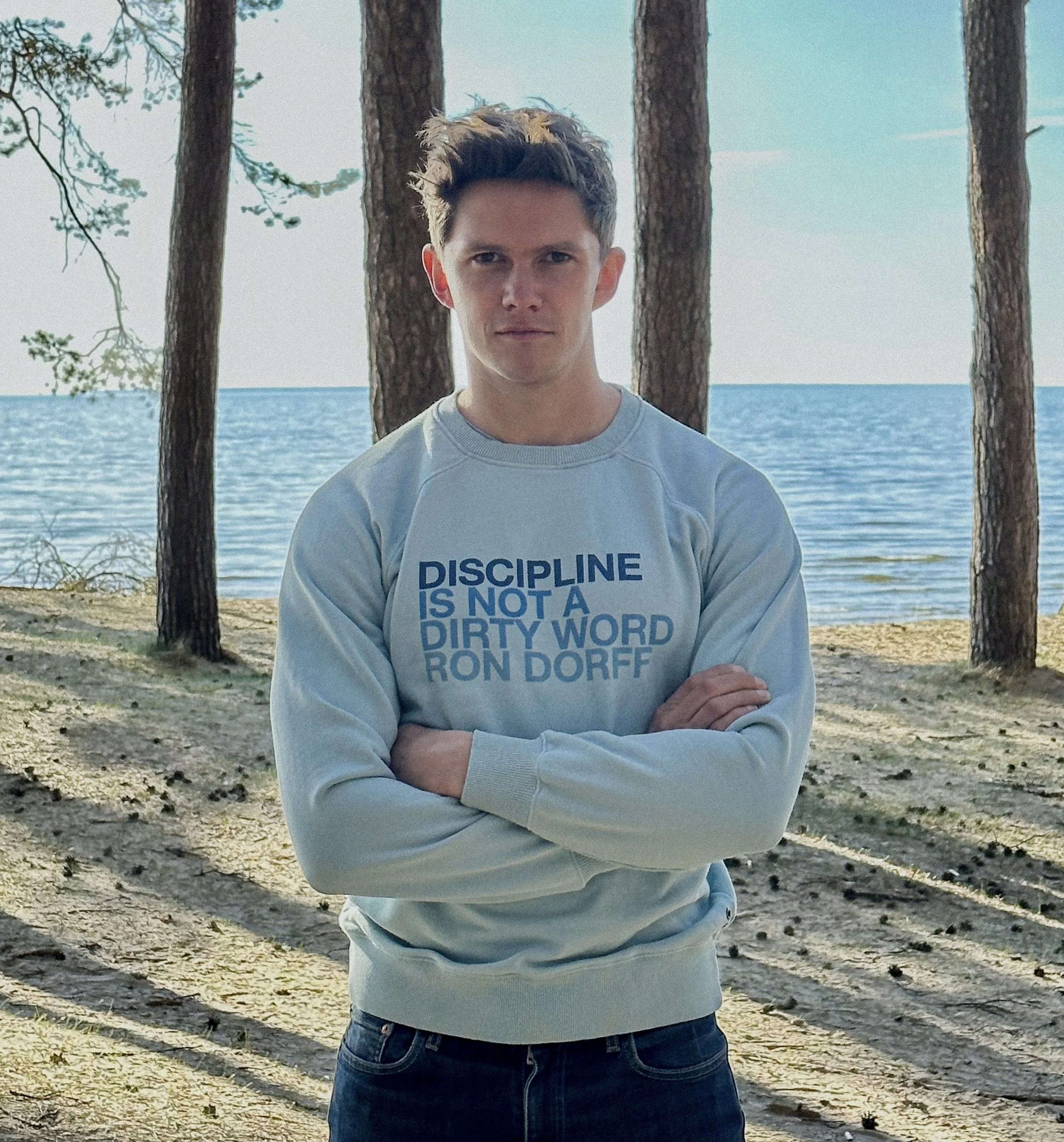 Tom Prior with a serious expression standing outdoors by a body of water with trees behind him, wearing a white sweatshirt with the message, 'Discipline is not a dirty word,' crossed arms.
