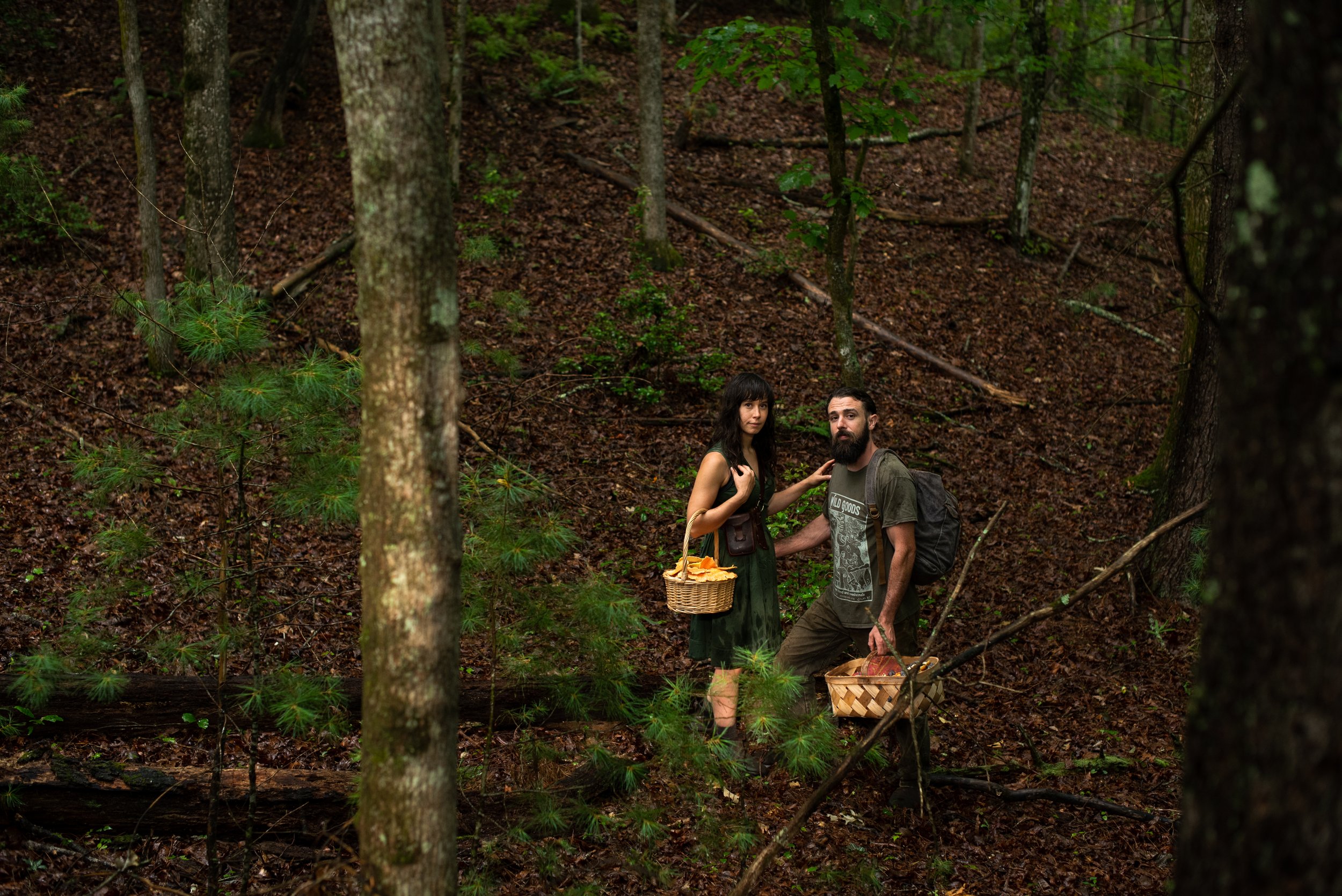 A man and woman standing in a forest, carrying baskets with leaves and mushrooms, surrounded by trees and fallen leaves.