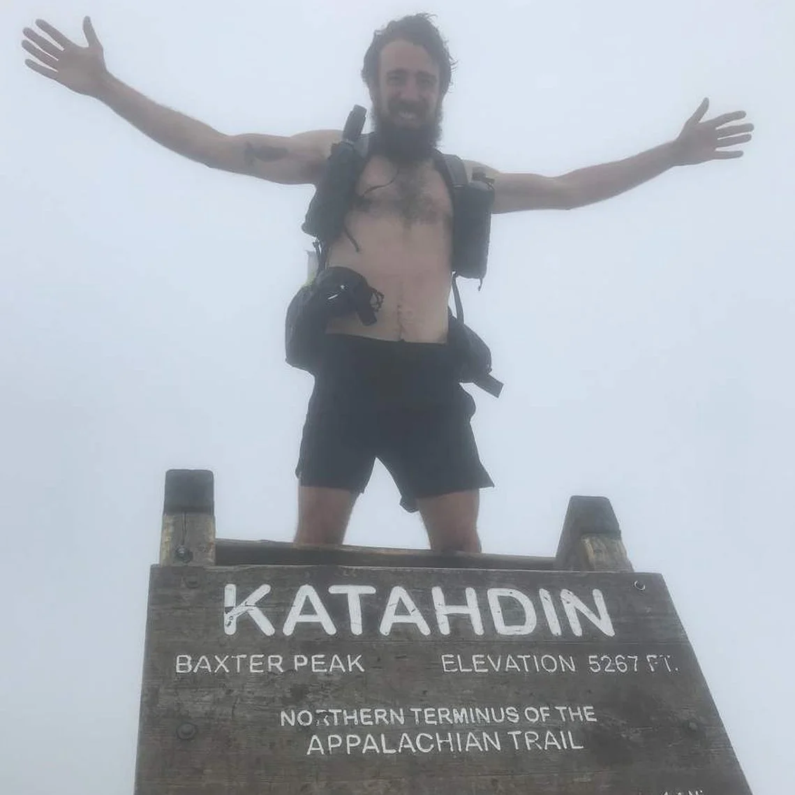 A man standing on a summit sign at Katahdin, Baxter Peak, with arms outstretched. He is shirtless, wearing black shorts, and carrying a backpack. The sign indicates an elevation of 5267 feet and marks the northern terminus of the Appalachian Trail, with the sky obscured by fog.
