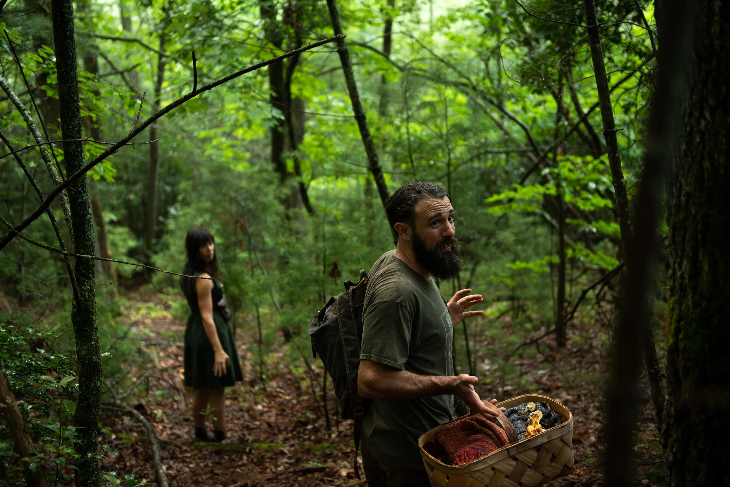 A man with a beard and long hair, carrying a backpack and holding a basket, standing on a forest trail and talking to a woman in a black dress, with trees and green foliage in the background.