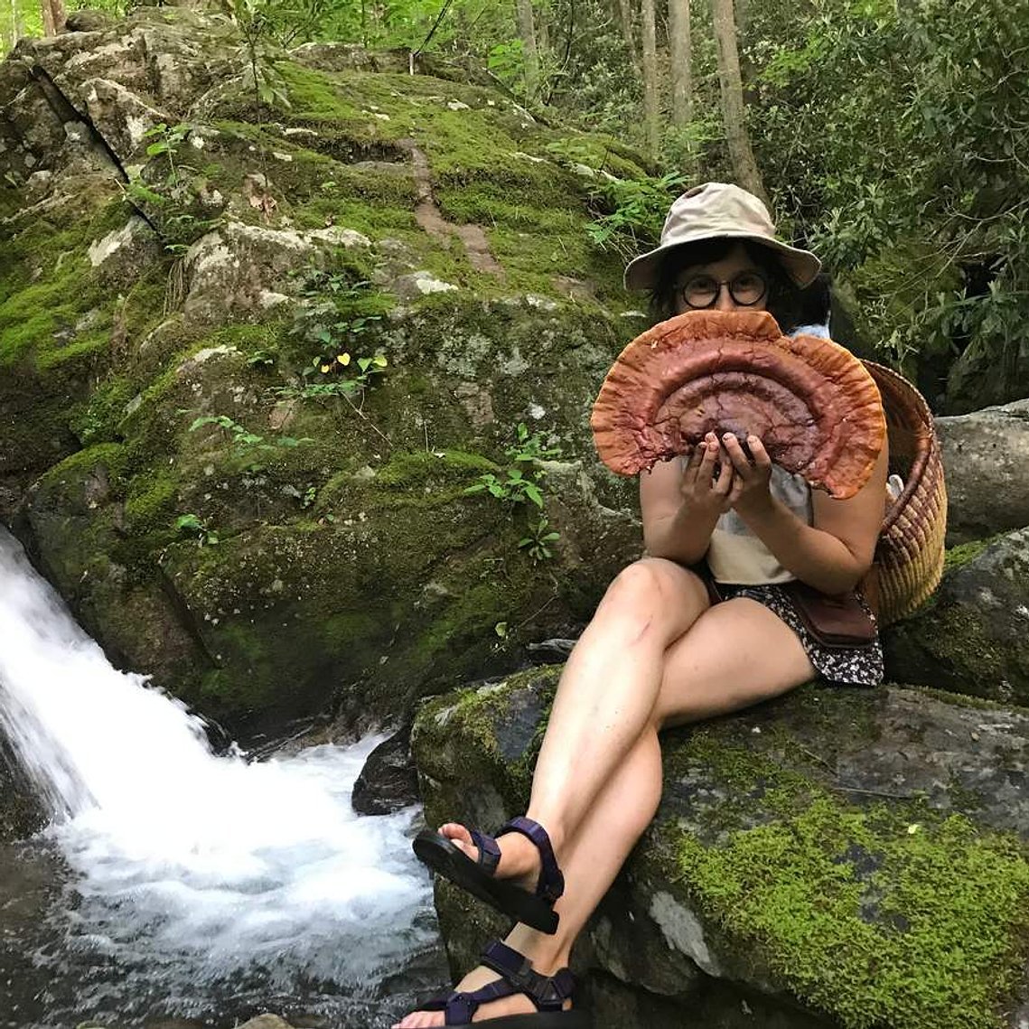 A woman sitting on moss-covered rocks beside a stream in a forest, holding a large mushroom and wearing a hat, glasses, and sandals.