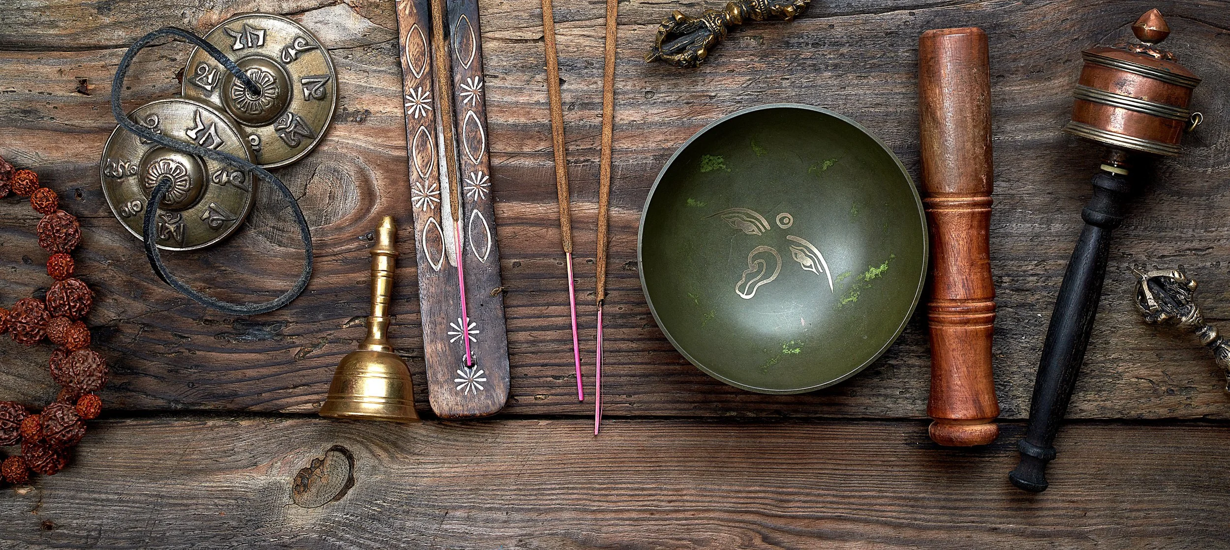 Assorted traditional Asian musical and prayer instruments on a wooden surface, including Tibetan tingsha cymbals, incense sticks, a singing bowl, and a prayer wheel.