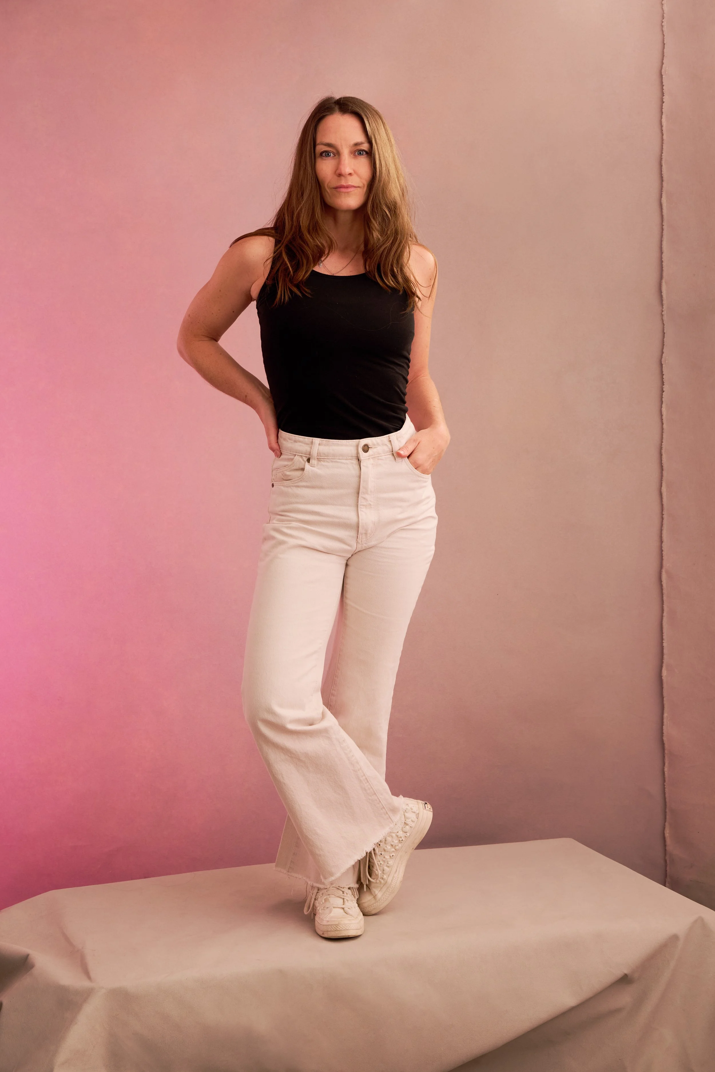 A woman with brown hair wearing a black sleeveless top, light-colored jeans, and white sneakers standing on a platform with a pink and beige background.
