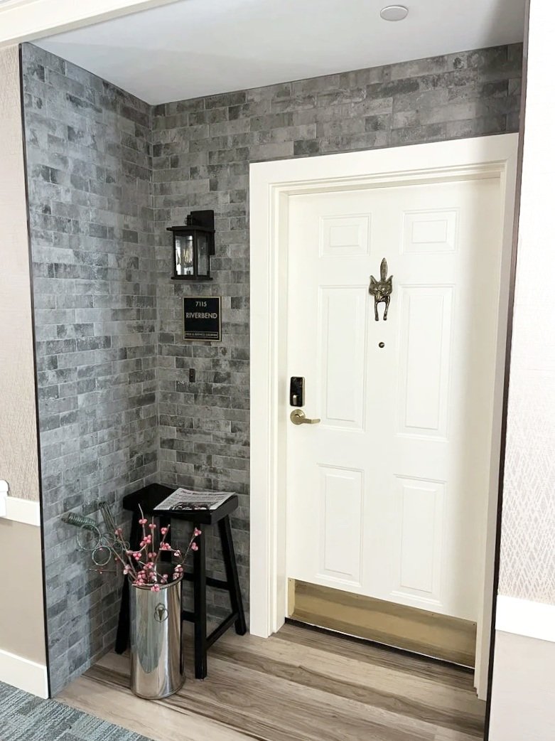 Entryway with brick wall, white door with gold handle, decorative donkey head, wall-mounted lantern, small black table, and metal container with pink flowers.
