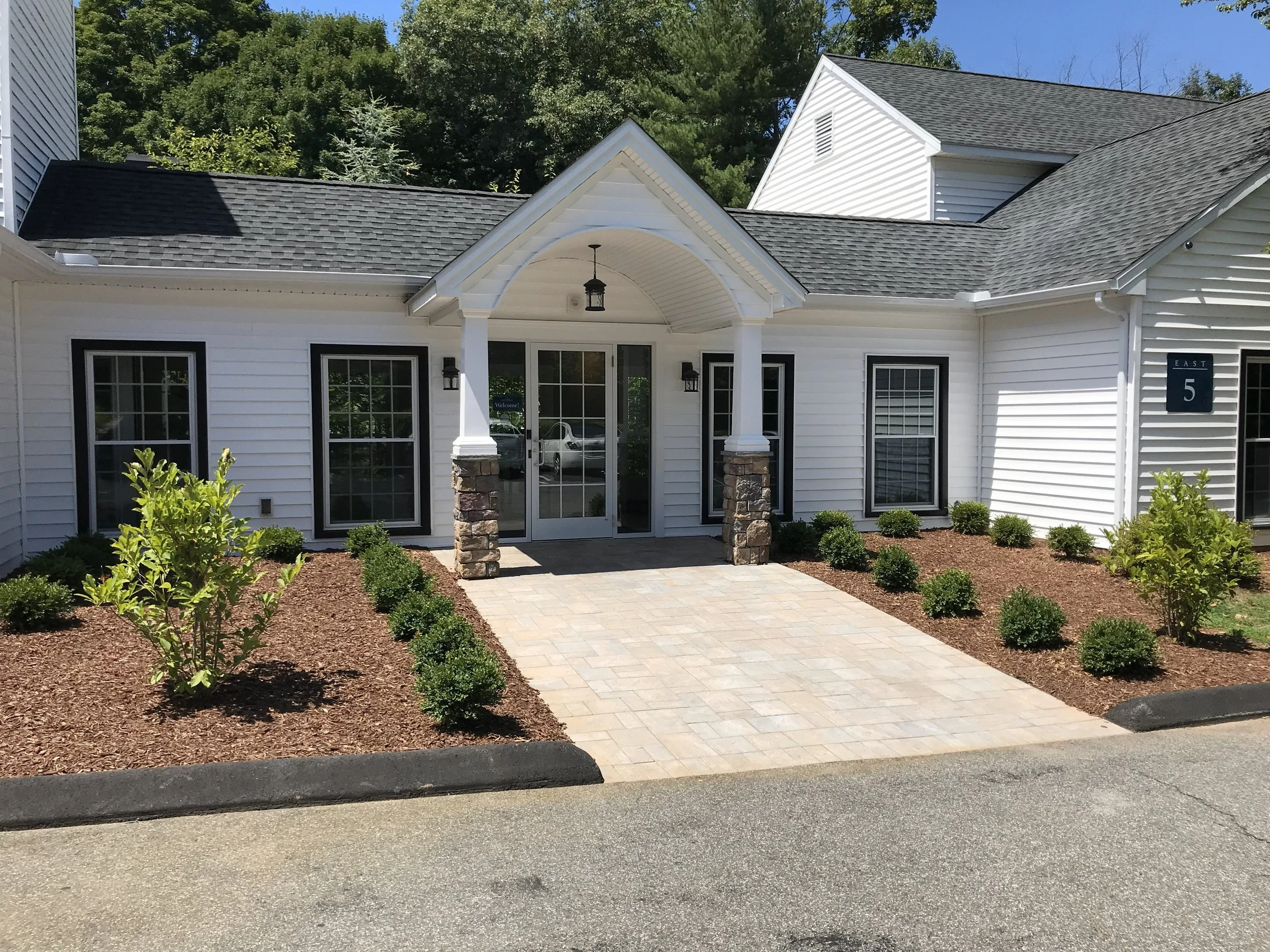 Front entrance of a white house with a stone and white column porch, four windows, landscaped garden beds, and a paved walkway.