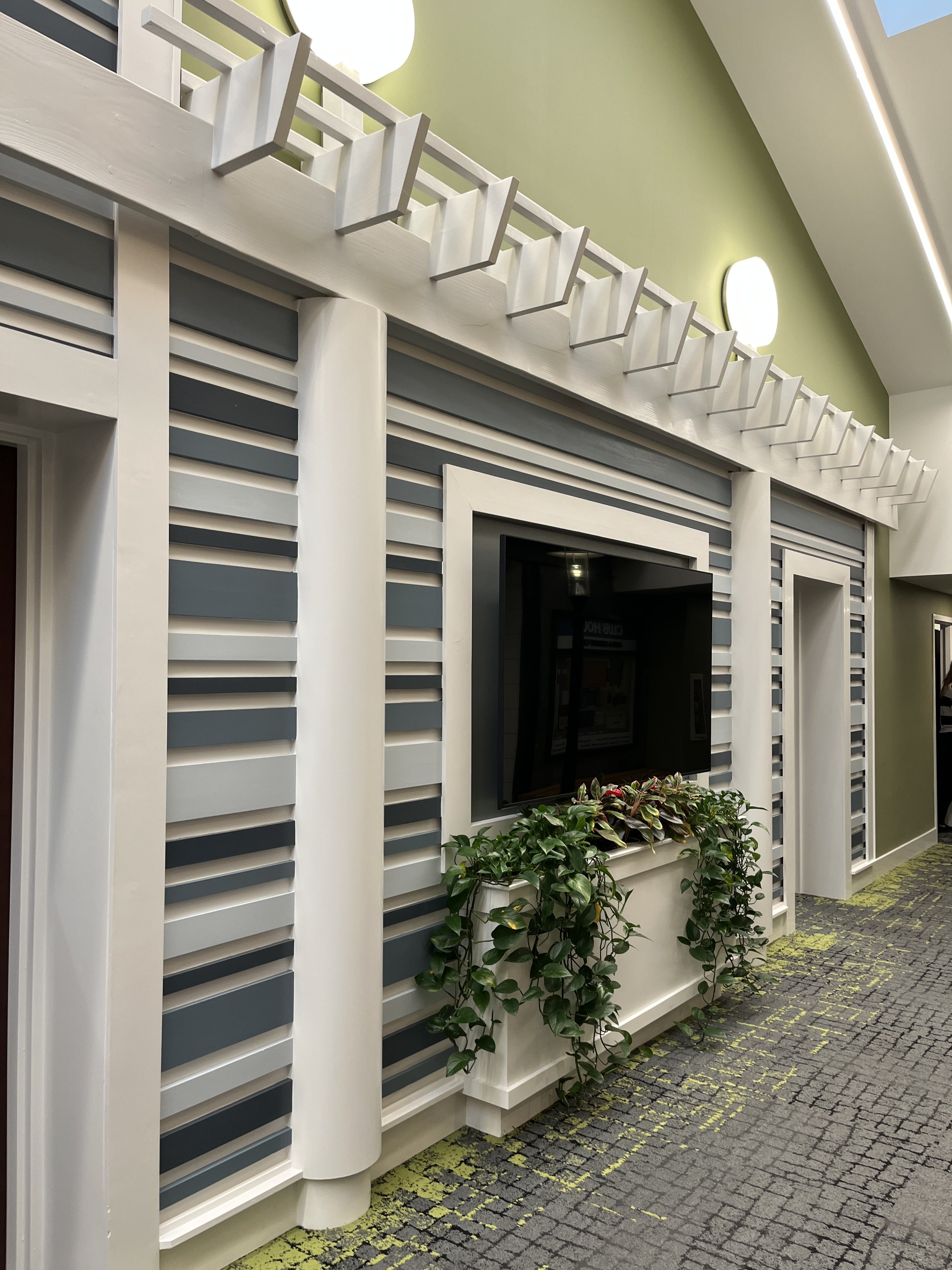 Modern interior wall with horizontal slats in gray and white, a mounted flat-screen TV, a plant in a white planter, and a green-colored wall with wall-mounted light fixtures.