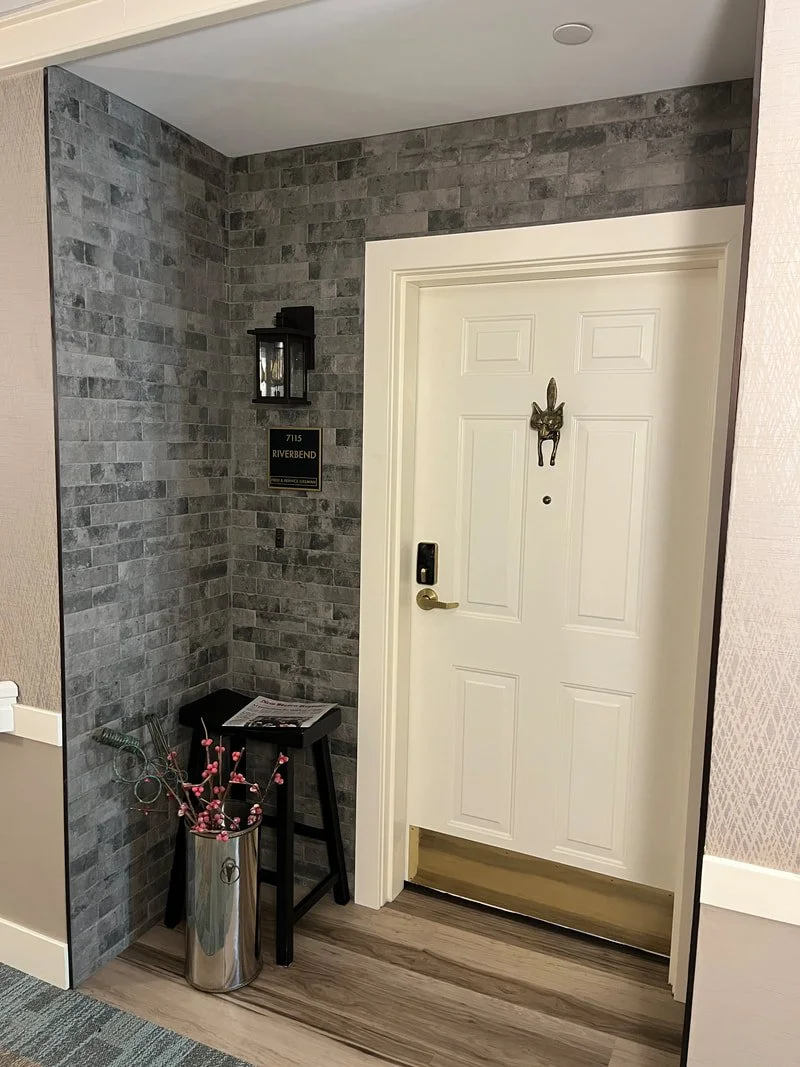 Interior entryway with a white door, gray brick accent wall, black wall-mounted lantern, and a small black stool with a brochure on top. A metallic vase with pink flowers is placed on the floor next to the stool.