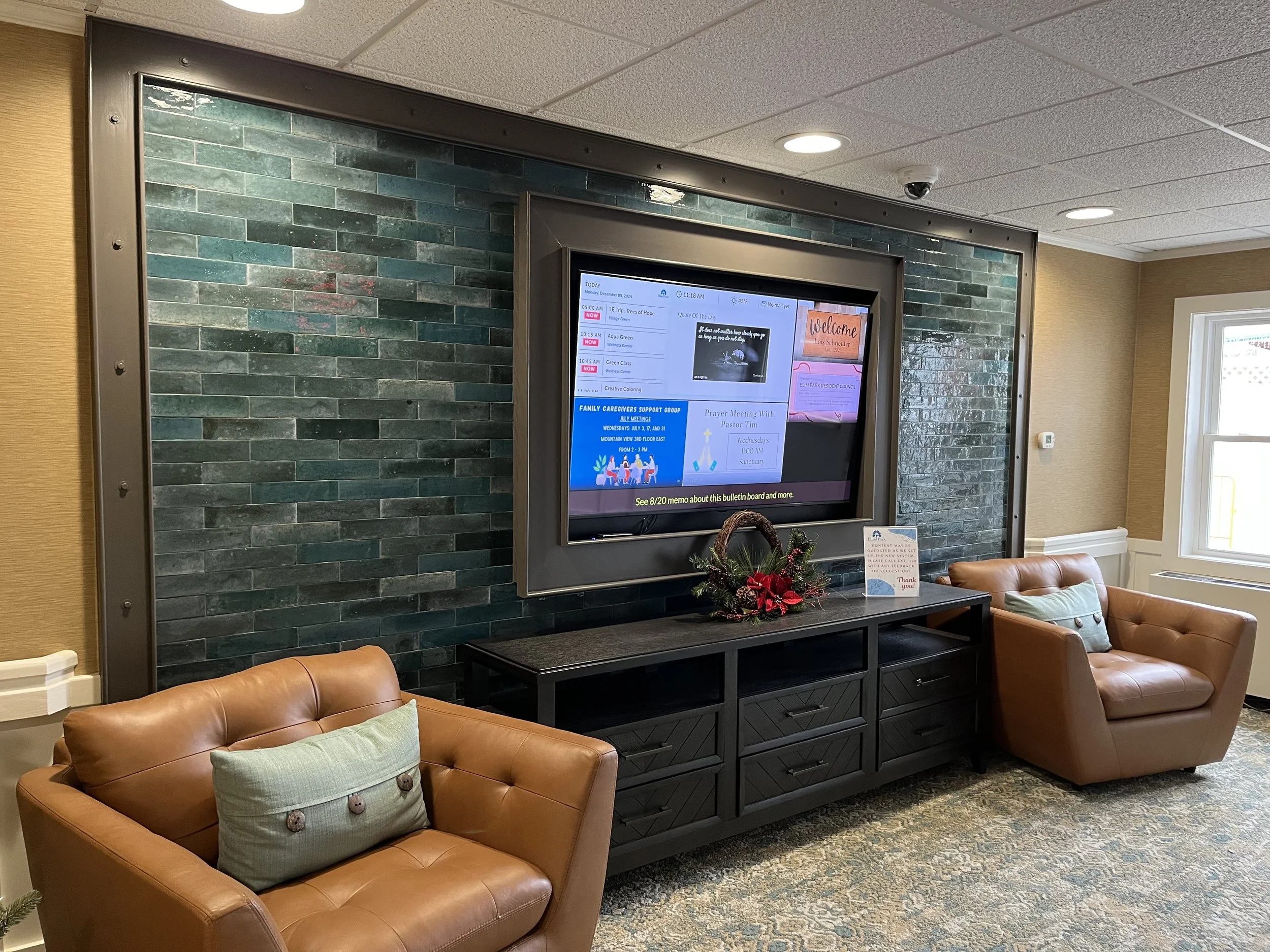 A waiting room with two tan leather chairs, a black entertainment console with holiday decorations, and a large screen displaying a bulletin board with schedules and notices on a blue tile wall.